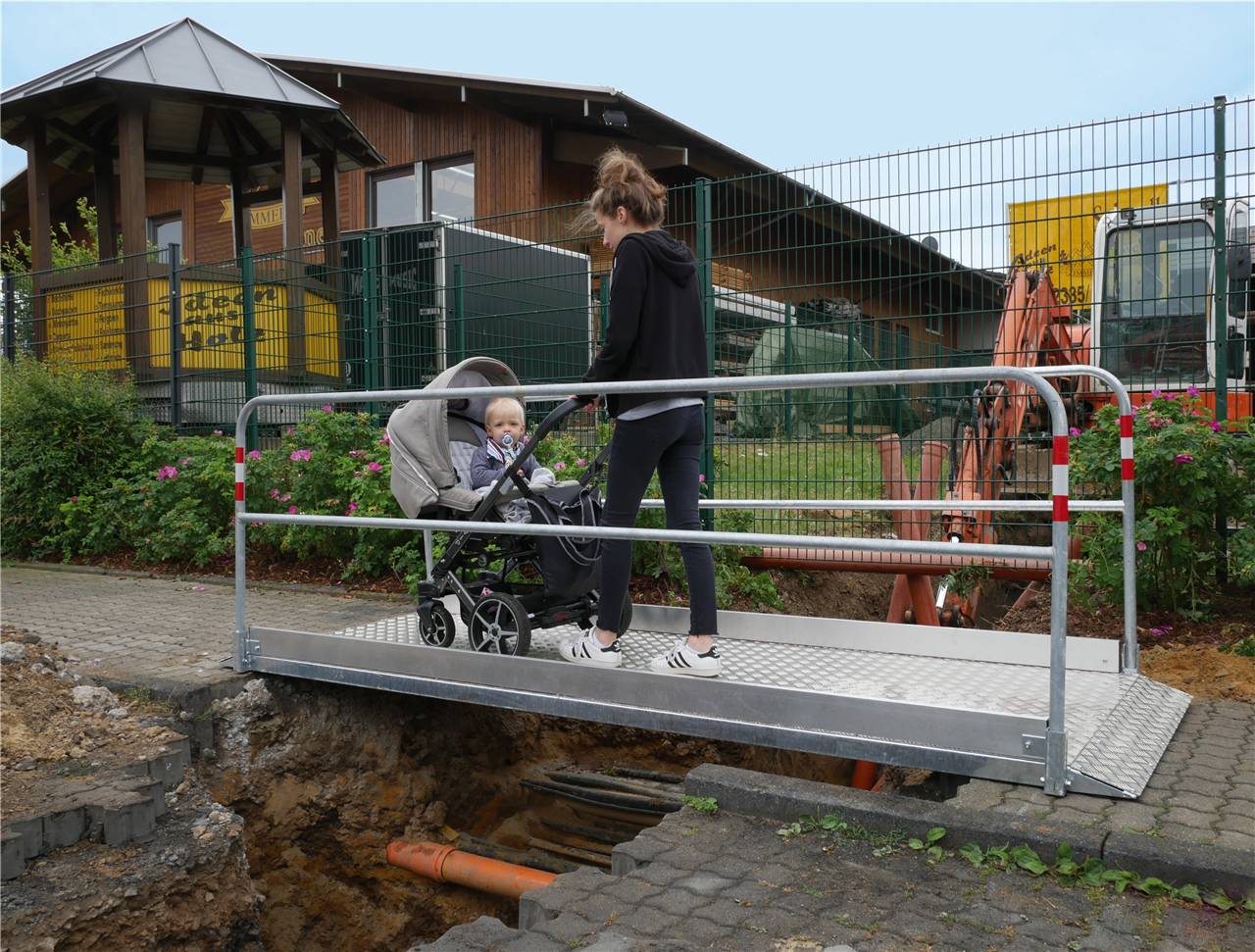 Eine Frau mit einem Kinderwagen überquert eine provisorische Metallbrücke über ein Baustellengebiet mit freigelegten Rohrleitungen, neben einem Holzgebäude.