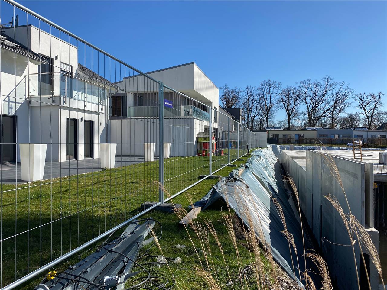 Eine Baustelle mit einem teilweise errichteten modernen Gebäude hinter einem Metallzaun. Aushubmaterialien und Werkzeuge sind im Vordergrund sichtbar.
