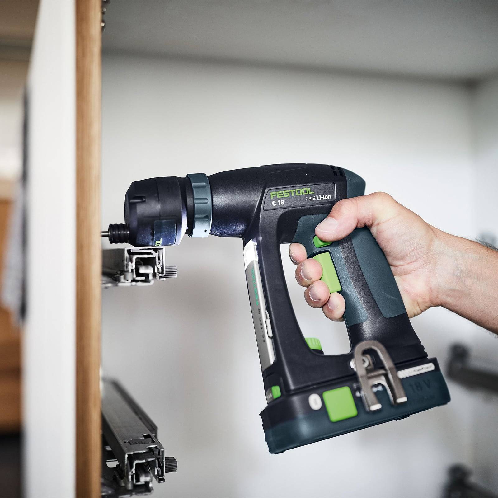 Eine Person hält einen kabellosen Akkuschrauber in der Nähe einer Holzoberfläche, wobei Metallschraubenbeschläge sichtbar sind. Der Akkuschrauber ist von der Marke 'Festool' und mit der Bezeichnung 'C 18 Li-ion' versehen.