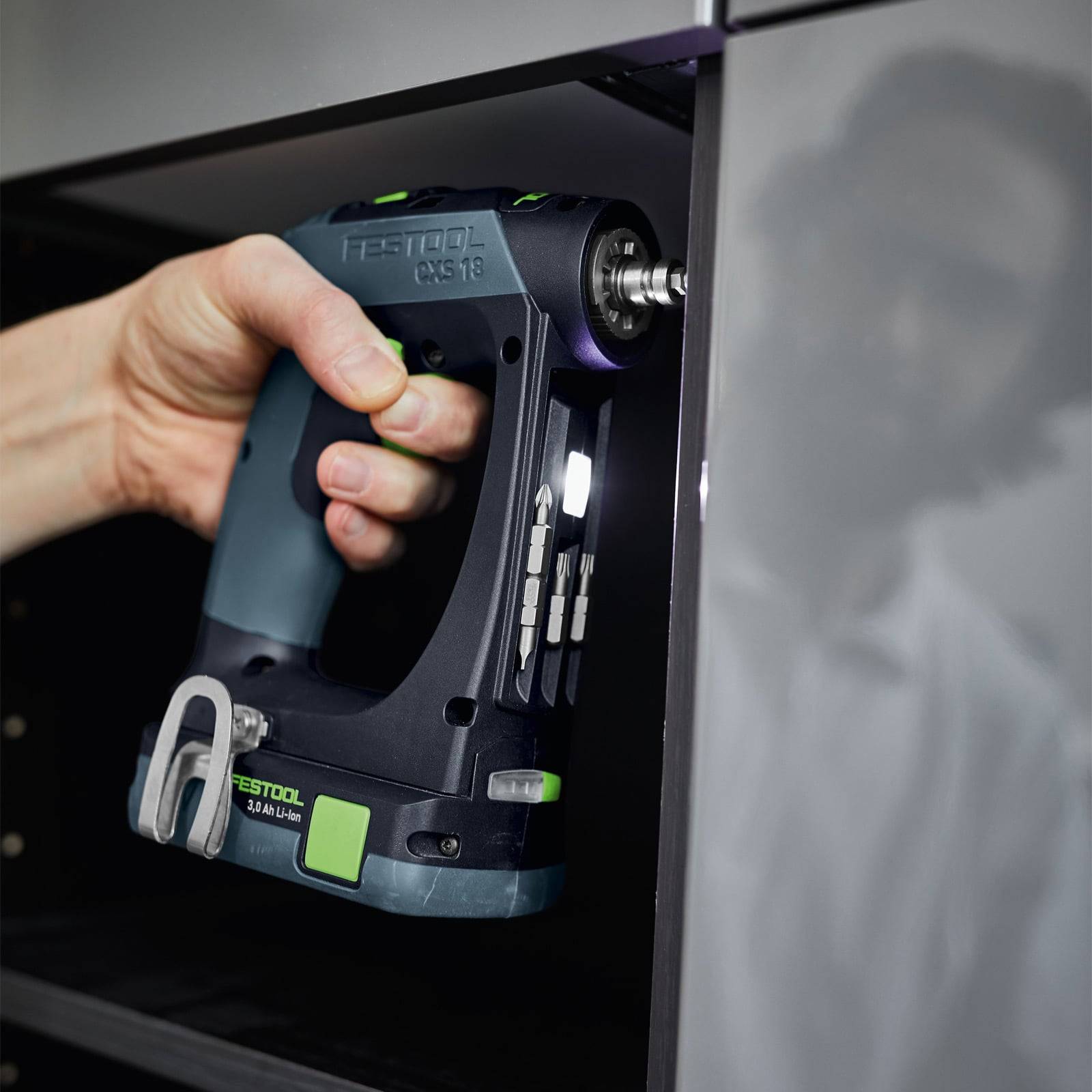 Eine Person verwendet einen tragbaren elektrischen Bohrer mit einem grün und schwarz gestalteten Gehäuse auf einer dunklen, glänzenden Oberfläche zum Festziehen oder Zusammenbauen.