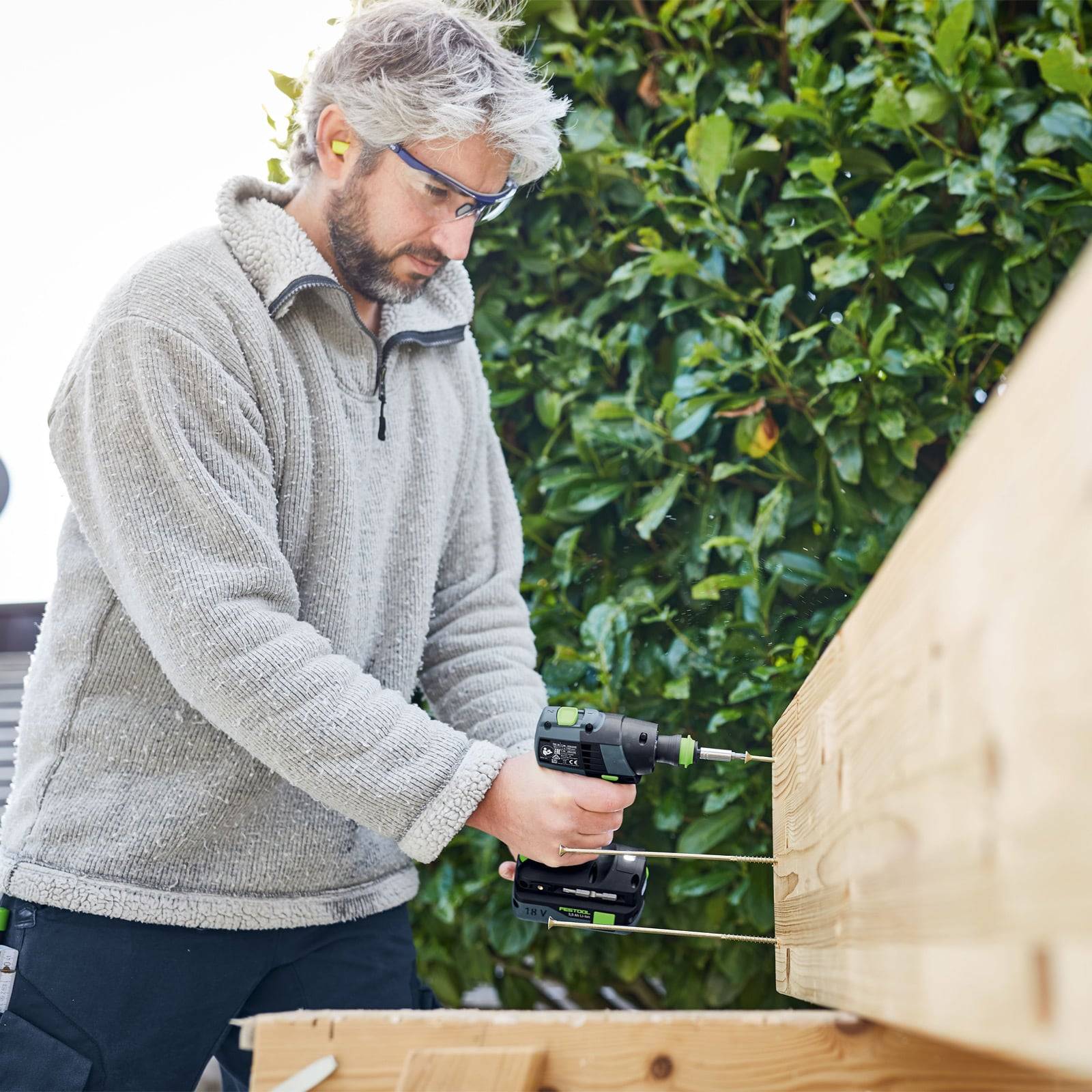 Eine Person mit Sicherheitsbrille verwendet einen Akkuschrauber, um Schrauben in Holzbalken im Freien zu treiben und steht dabei neben einer hohen Hecke.