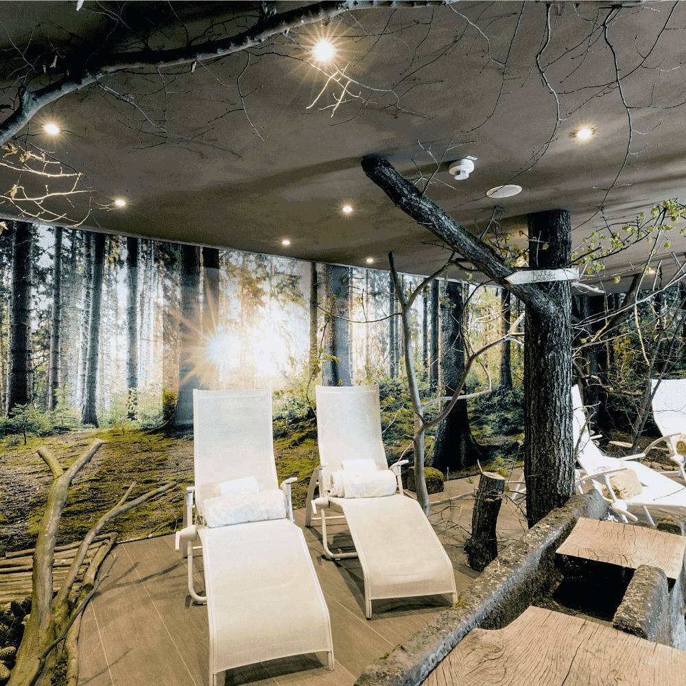 Entspannungsbereich eines Indoor-Spas mit weißen Liegestühlen und einem Waldgemälde, umgeben von baumähnlicher Dekoration und sanften Deckenleuchten.