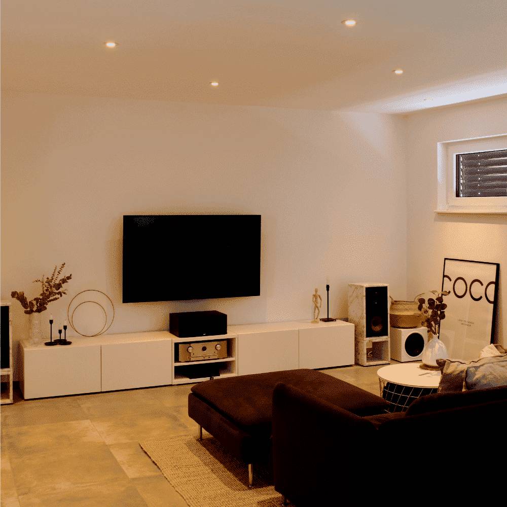 Ein moderner Wohnraum mit einer wandmontierten TV, einem weißen Schrank, Lautsprechern und minimaler Dekoration, mit einem Fenster, dessen Jalousien teilweise geschlossen sind.