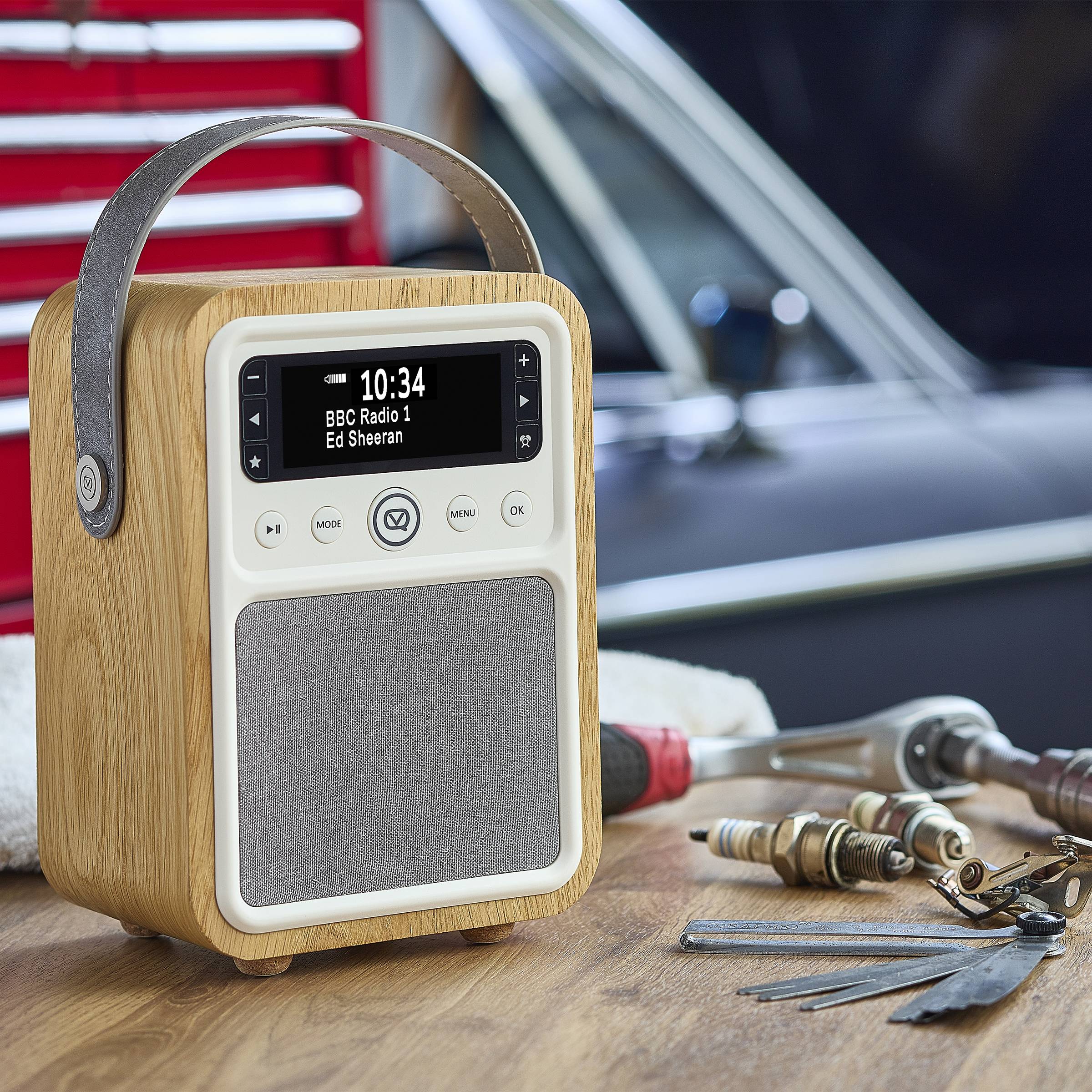 Ein Radio im Vintage-Stil auf einem Holztisch in einer Garagenumgebung zeigt auf seinem Bildschirm '10:34', 'BBC Radio 1' und 'Ed Sheeran' an.