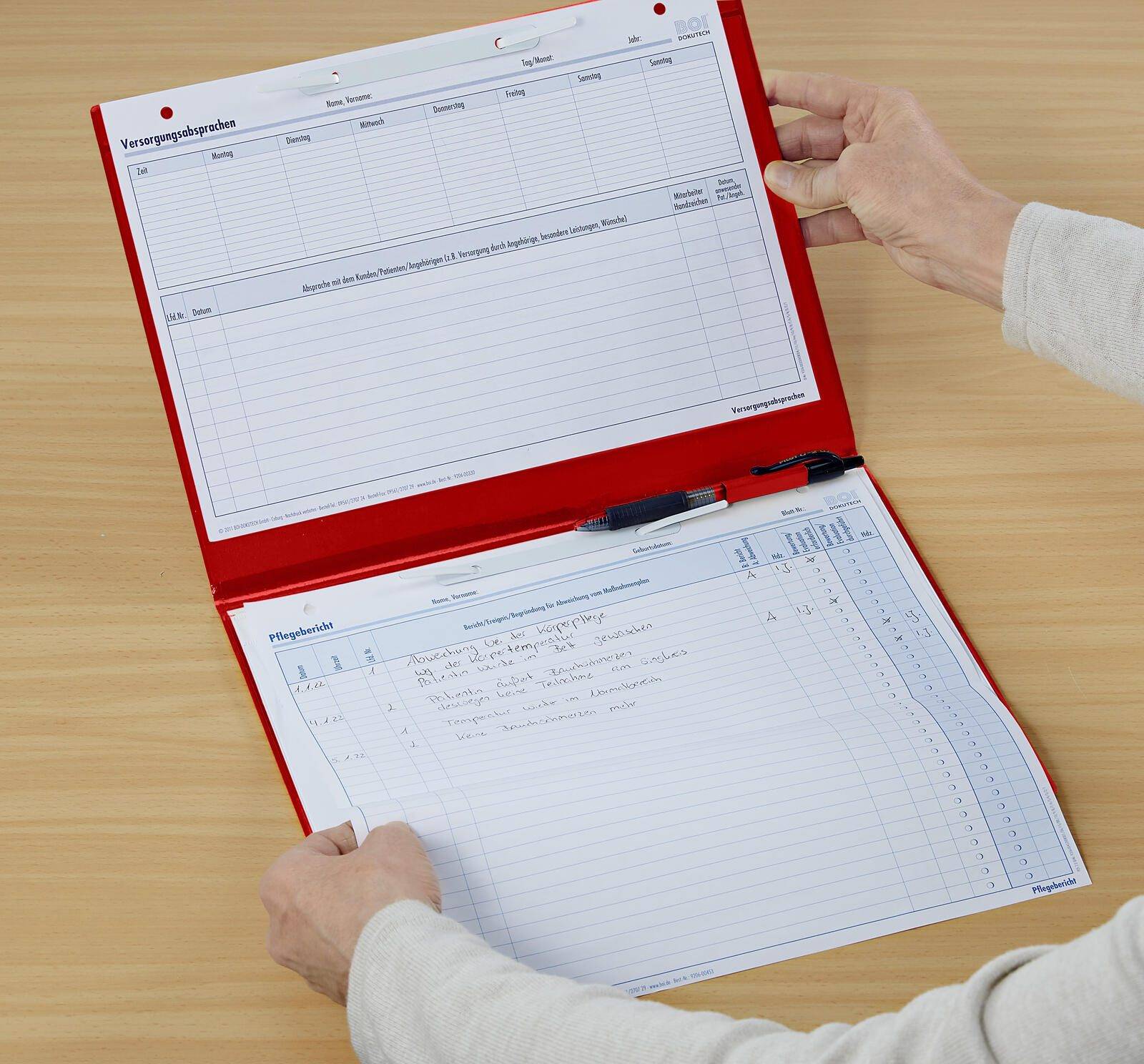 Eine Person öffnet eine rote Klemmbrett mit Blättern von Dokumenten darin, möglicherweise ein Formular oder eine Checkliste, auf einer hellen Holzoberfläche.
