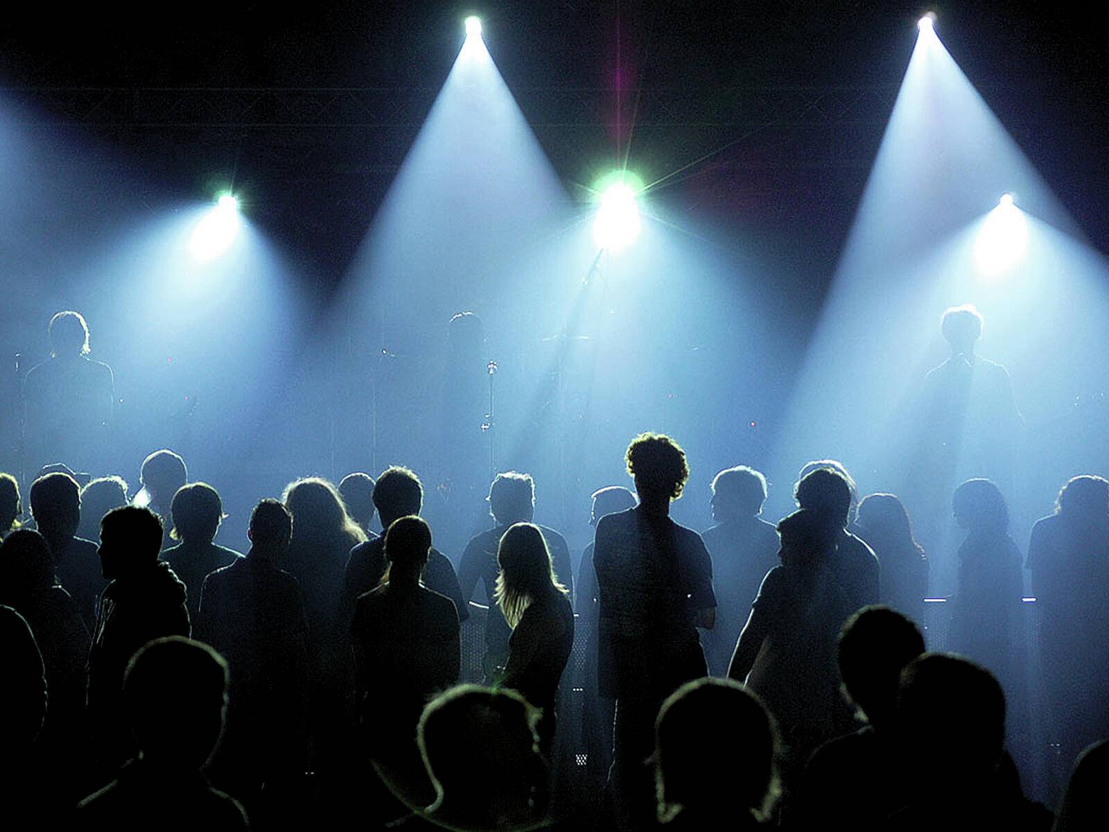 Publikum als Silhouetten vor einer hellen Bühne mit mehreren Scheinwerfern, die eine lebendige Konzatatmosphäre erzeugen.