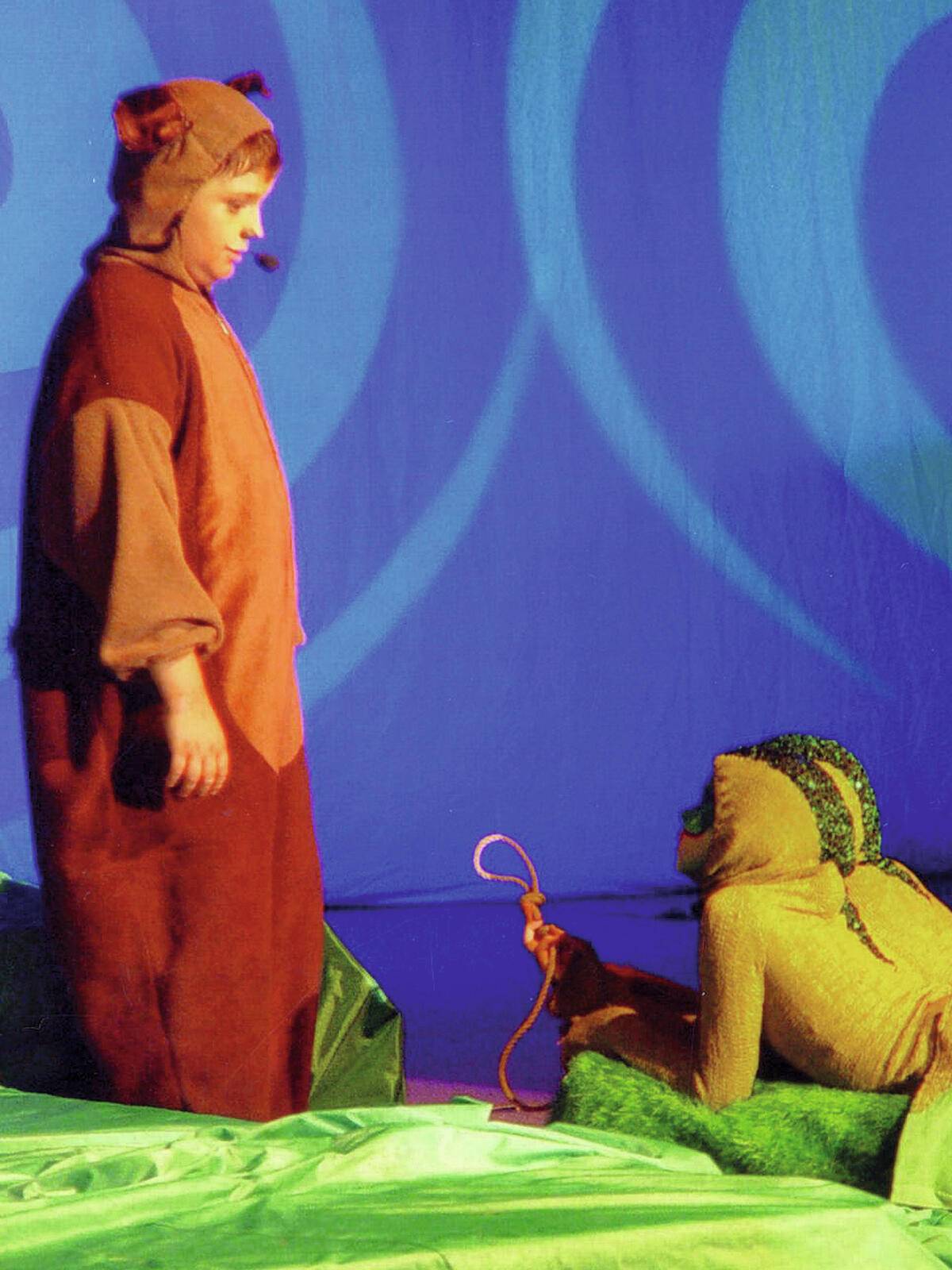 Zwei Menschen in Tierkostümen, einer stehend und einer hockend, treten auf einer Bühne mit buntem Hintergrund auf, offensichtlich in einer Theaterszene.