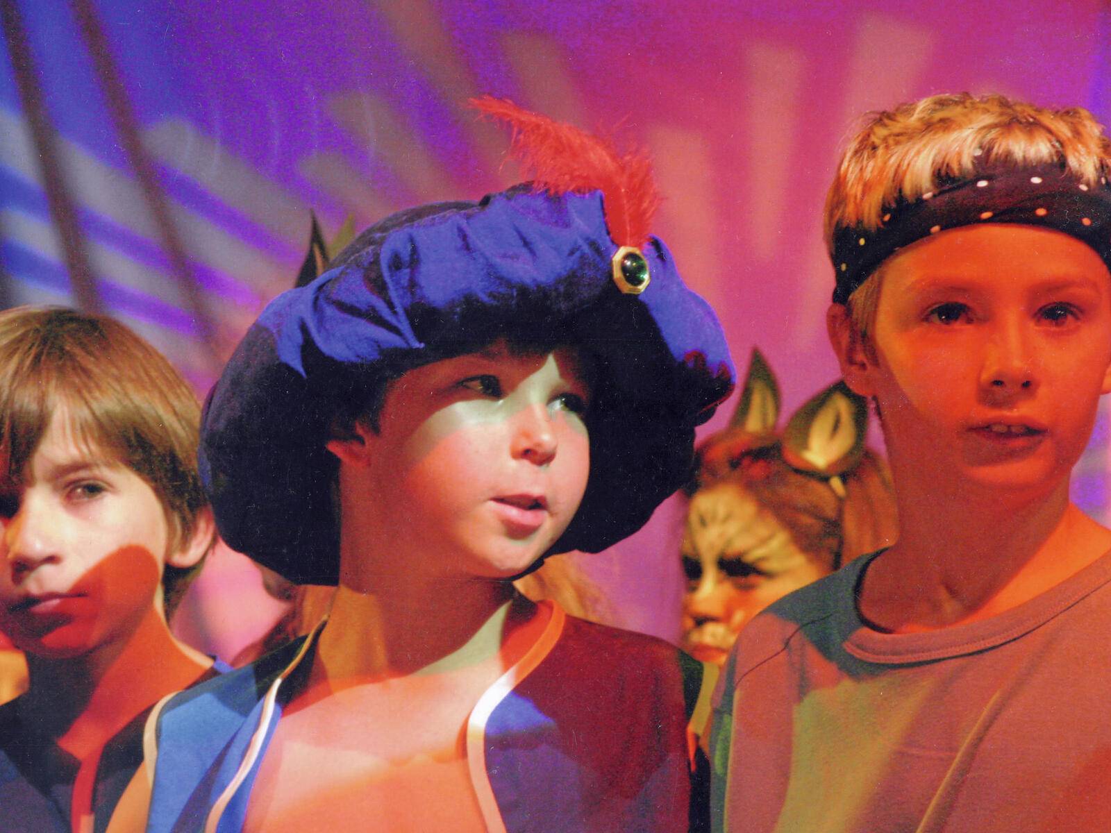 Kinder in bunten Kostümen, darunter ein blauer Hut mit einer Feder, nehmen an einer Aufführung auf einer Bühne teil, die mit violettem Licht beleuchtet ist.