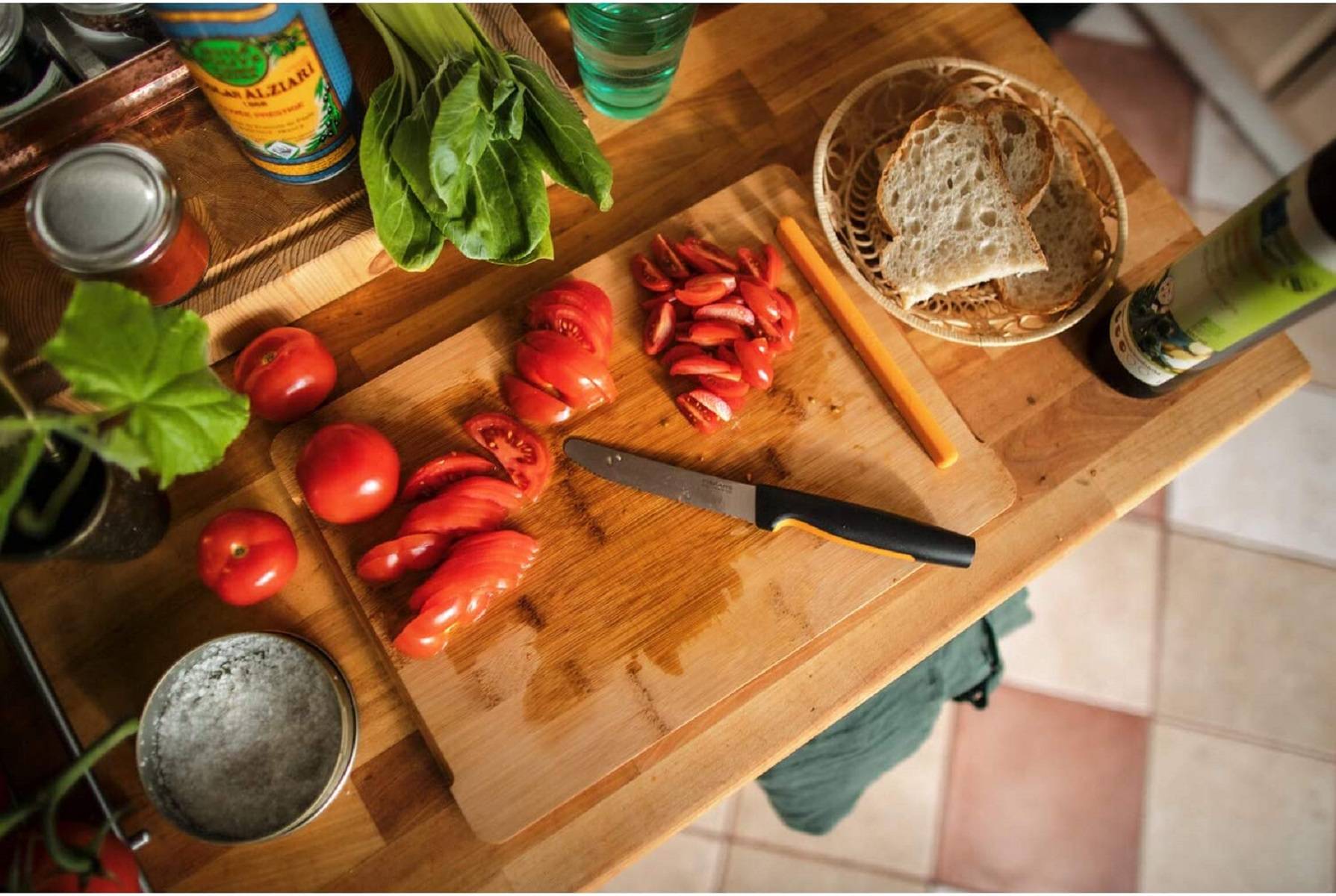 Draufsicht auf ein Schneidbrett in einer Küche mit geschnittenen Tomaten, Basilikum, einem Messer, Brot in einem Korb und Speisekammer-Artikeln zum Kochen.