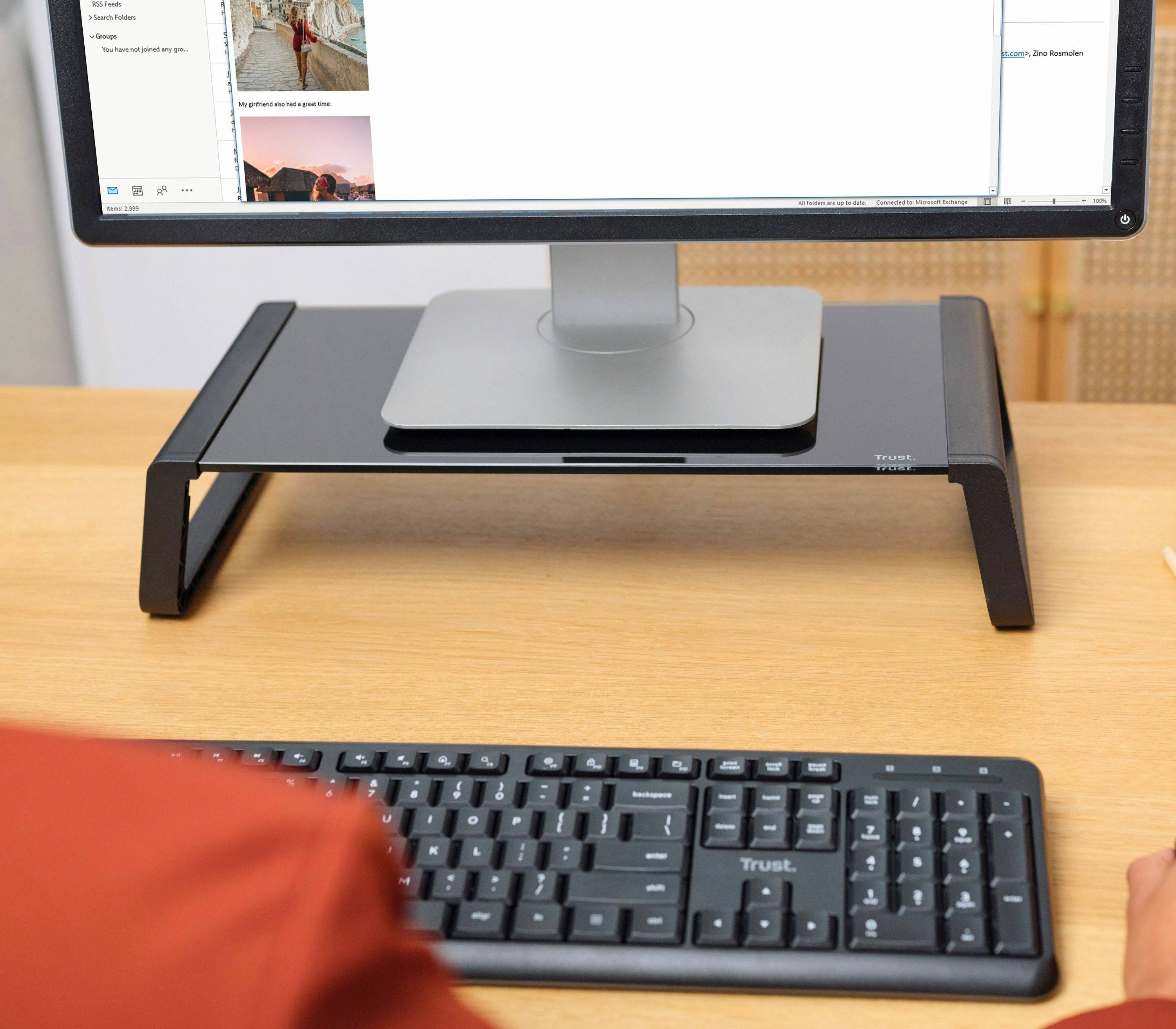 Desktopmonitor auf einer schwarzen Standerhöhung über einer Tastatur auf einem Holzschreibtisch, mit einer Person, die am Computer sitzt.