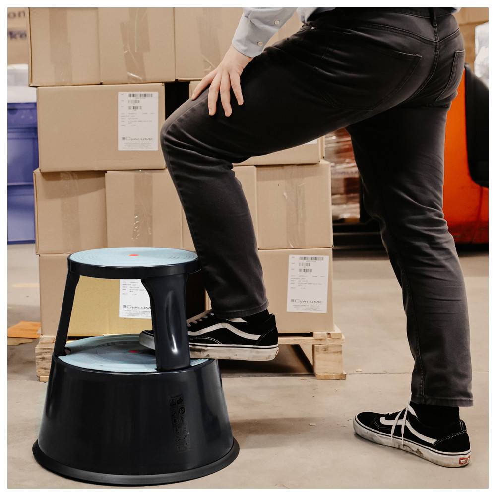Eine Person tritt auf einen kleinen schwarzen Hocker in einem Lagerhaus und stabilisiert einen Stapel Kartonboxen auf einer Holzpalette.