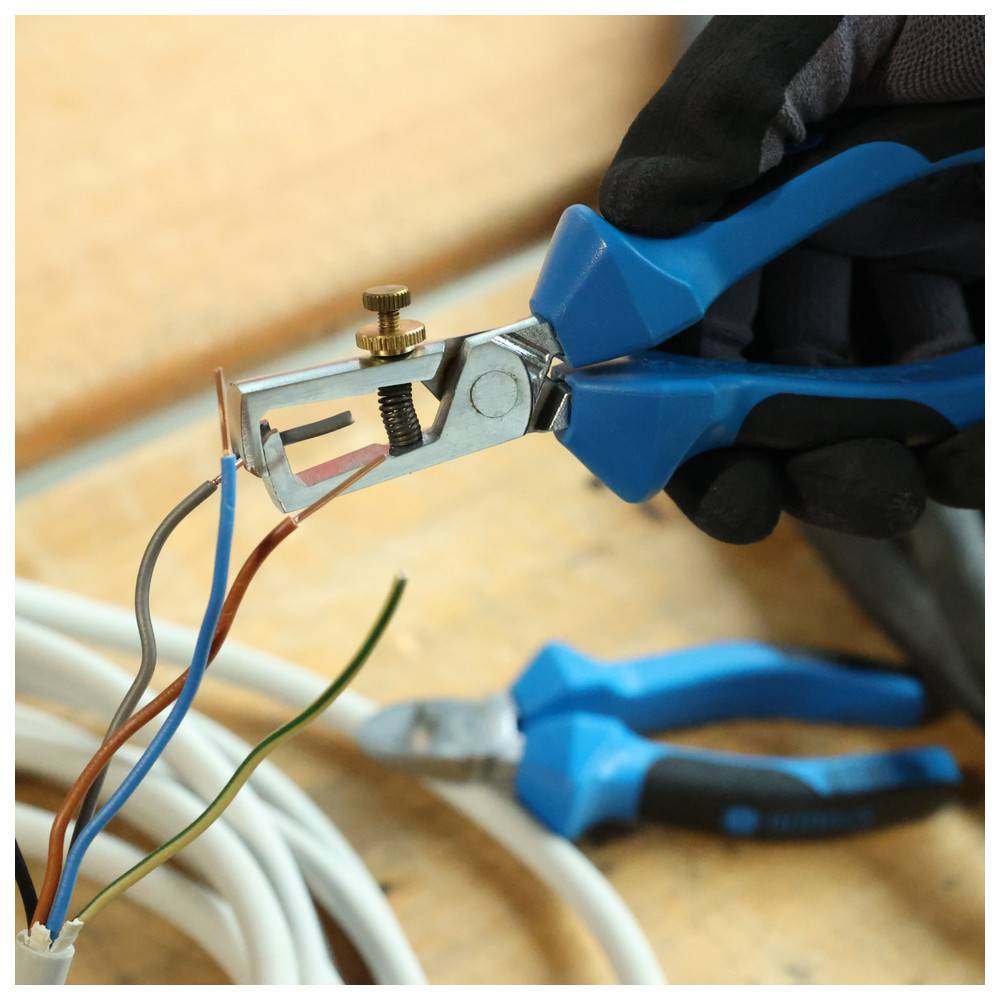 Eine Hand, die Abisolierzangen verwendet, um die Isolierung von elektrischen Kabeln zu entfernen, mit Zangen im Hintergrund auf einer Holzoberfläche.