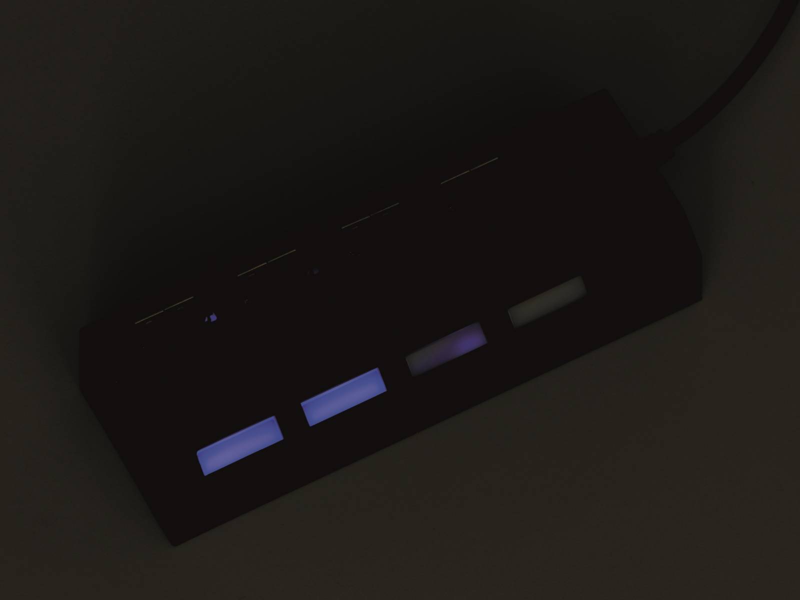 Ein dunkler Raum zeigt ein von hinten beleuchtetes Bedienfeld mit vier leuchtenden Tasten, drei in Lila und eine in Weiß, was auf ein elektronisches Gerät hindeutet.