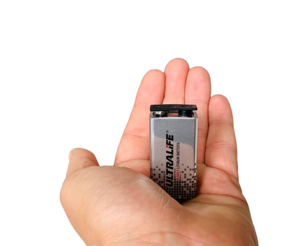 Hand, die eine kleine silberne Ultralife-Batterie gegen einen schlichten schwarzen Hintergrund hält.