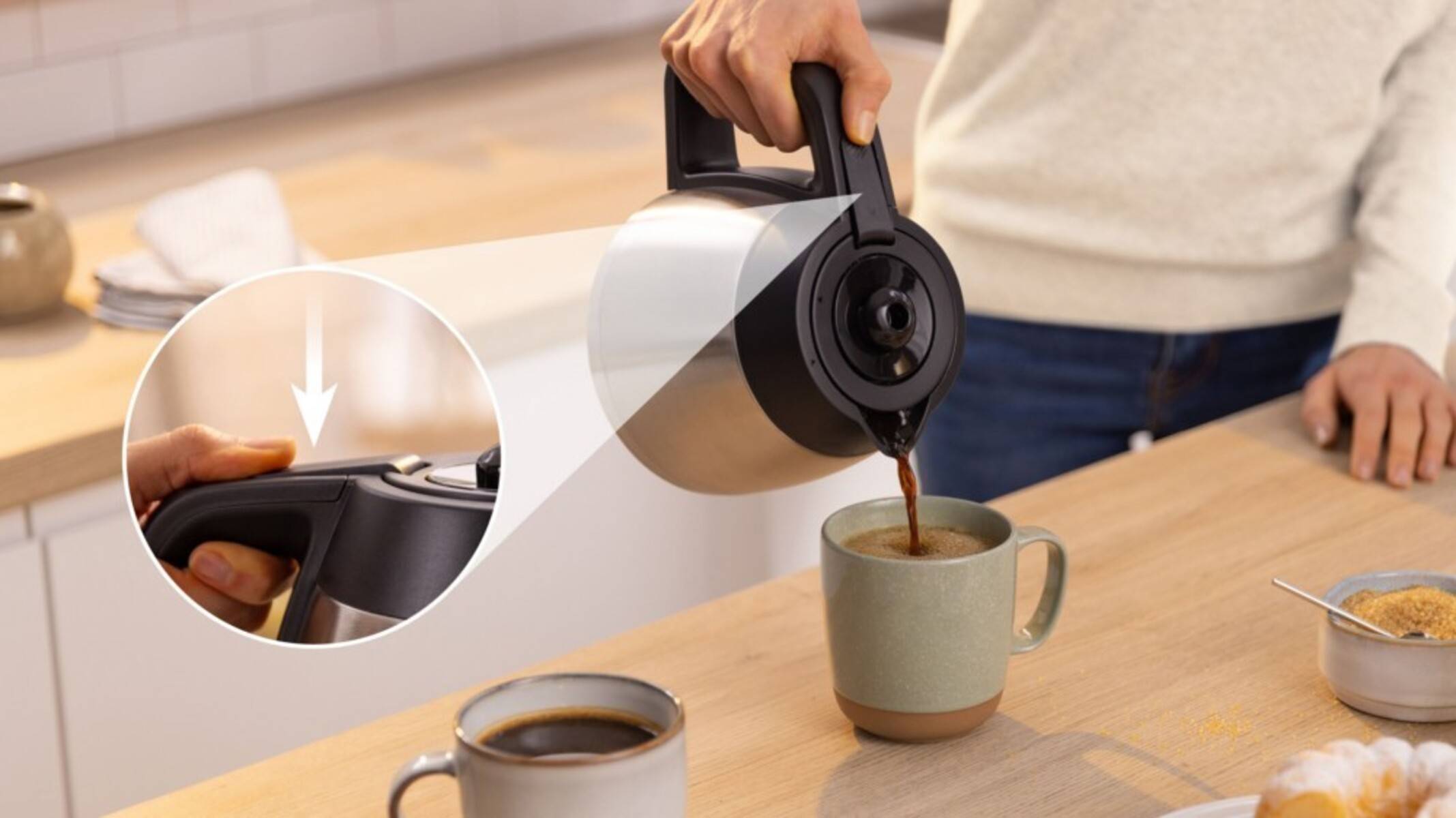 Eine Person gießt Kaffee aus einer Karaffe in eine Keramiktasse und betont dabei das Ausgießmerkmal. Eine zweite Tasse steht auf dem Holztisch.