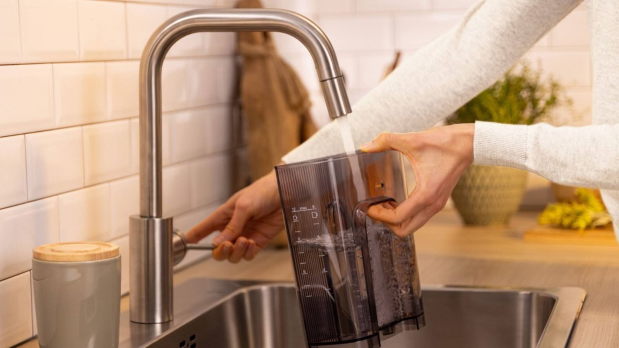 Eine Hand, die einen transparenten, markierten Wasserkrug an einem modernen Wasserhahn in der Küche auffüllt, mit einer gefliesten Rückwand im Hintergrund.
