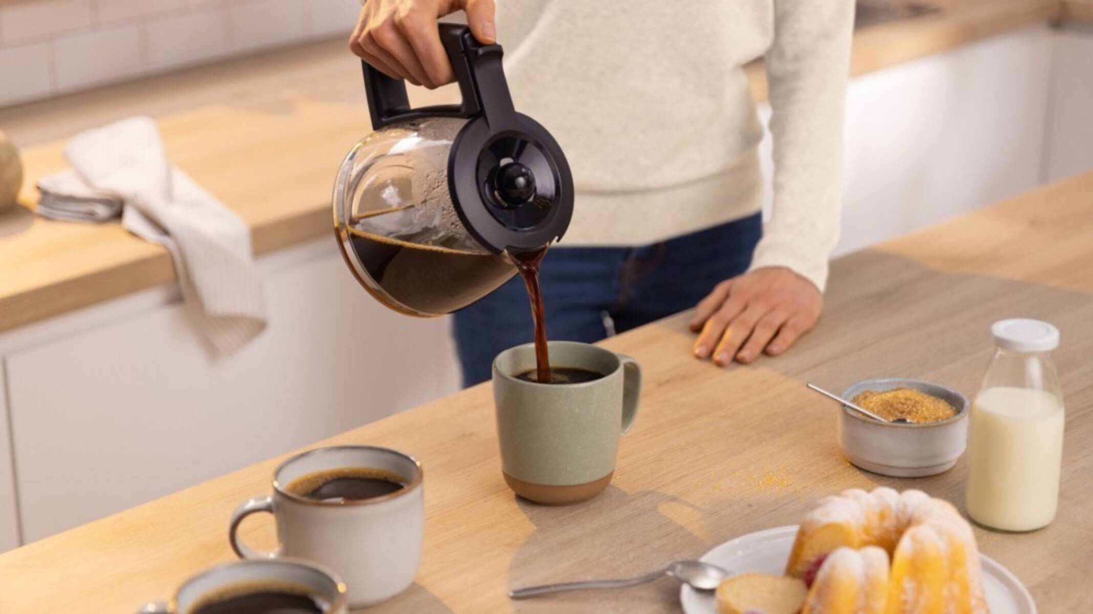 Eine Person gießt Kaffee aus einer Karaffe mit schwarzem Griff in einen grünen Becher auf einer Holzküche-Arbeitsfläche, mit zwei weiteren Bechern in der Nähe.