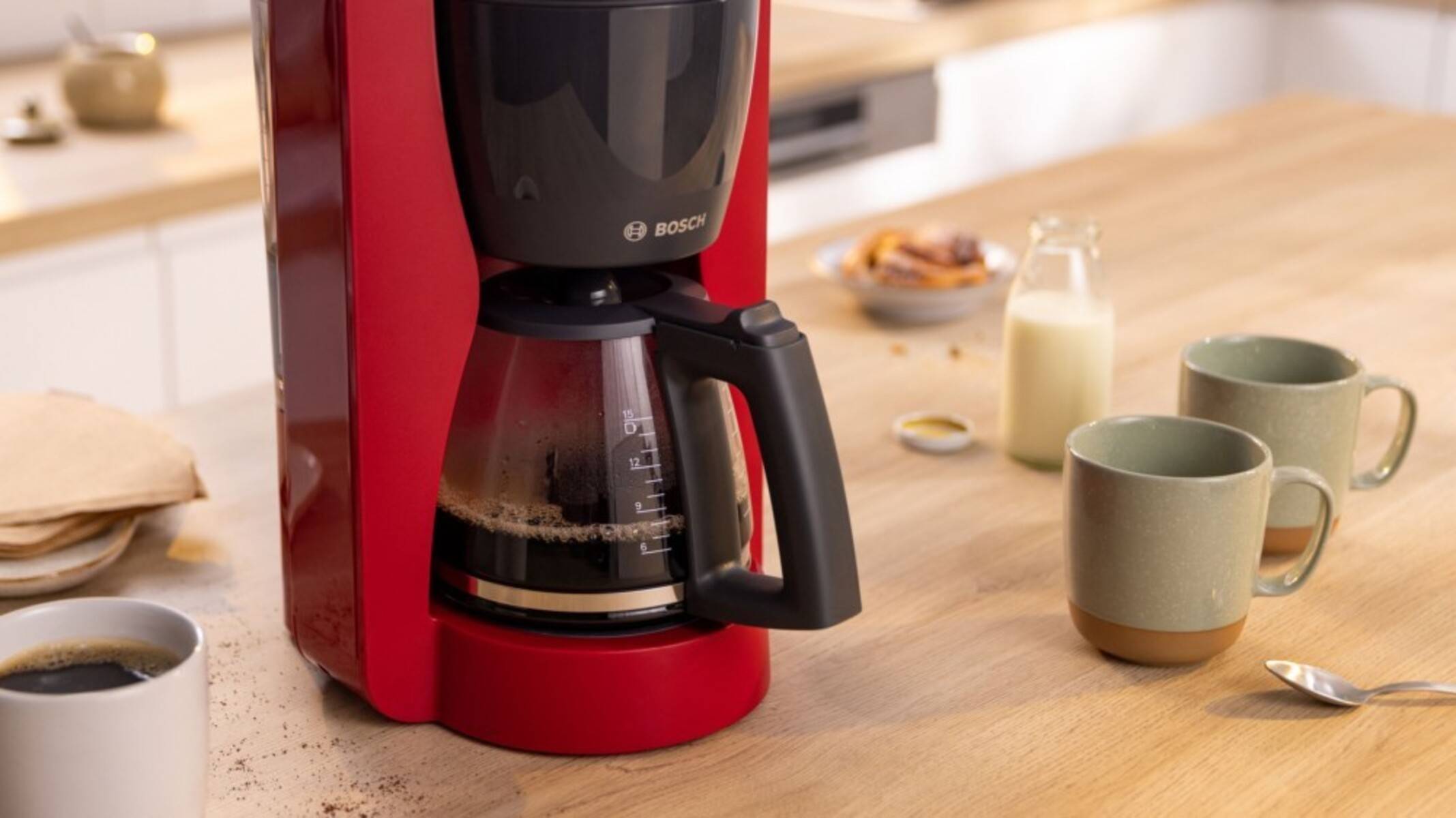 Rote Kaffeemaschine, die Kaffee in eine Glaskanne auf einem Küchentisch braut, mit zwei Bechern, einer Milchflasche und einer Kaffeetasse in der Nähe.