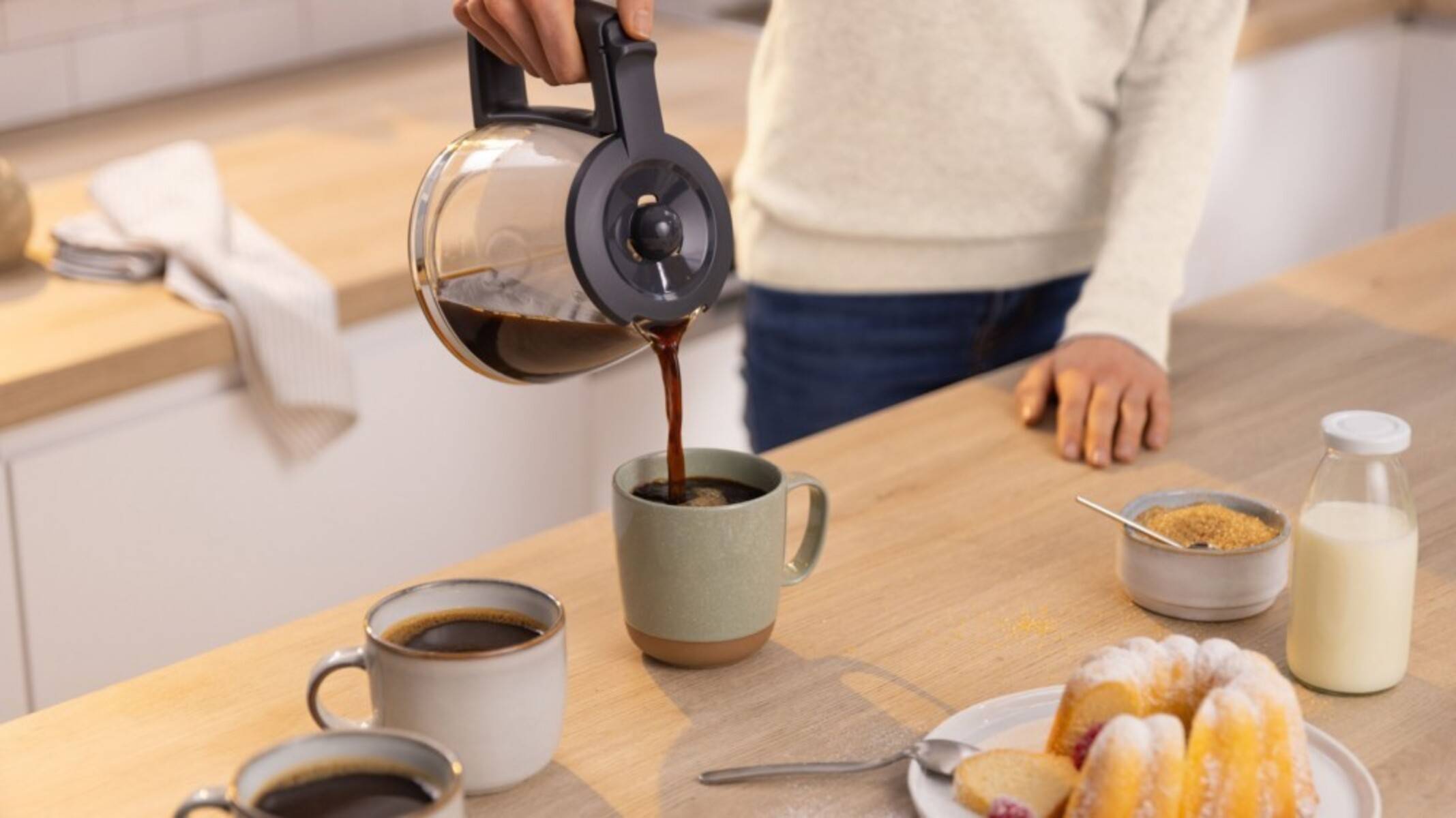 Eine Person gießt Kaffee aus einer Glaskanne in eine Tasse an einem Küchentisch mit anderen Kaffeetassen, Brot, Milch und einer Schüssel Müsli.