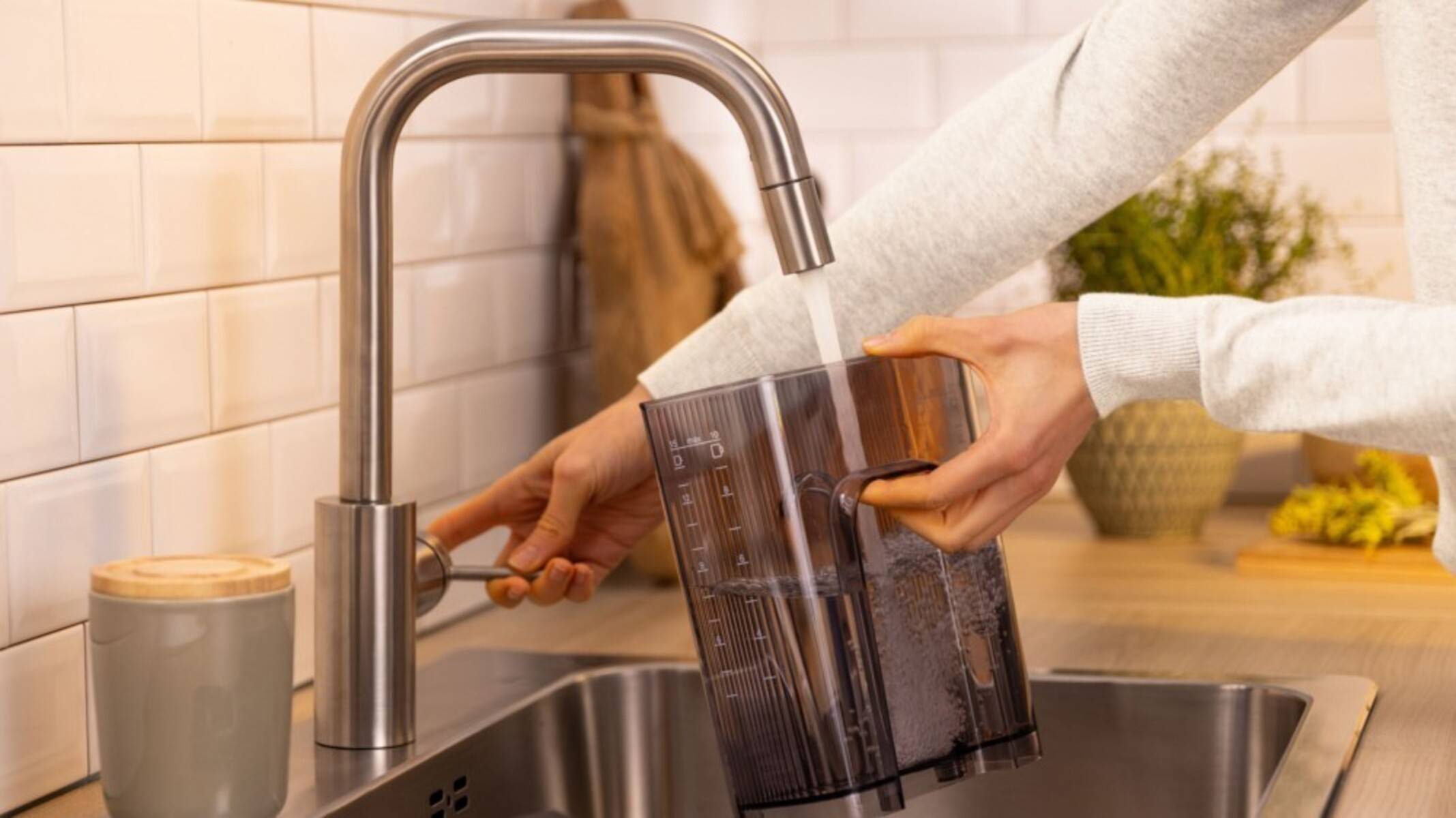 Eine Person füllt einen durchsichtigen Wasserkrug von einem modernen silbernen Wasserhahn in der Küche, wobei die Handlung des Wasserschöpfens in einem häuslichen Umfeld hervorgehoben wird.