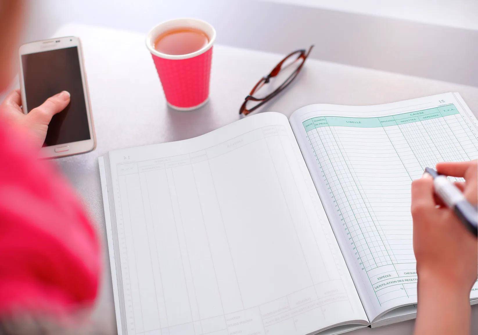 Offenes Ledger mit sichtbaren Spalten, eine Brille, eine rote Kaffeetasse und ein Tablet, alles auf einem Tisch in heller Farbe.