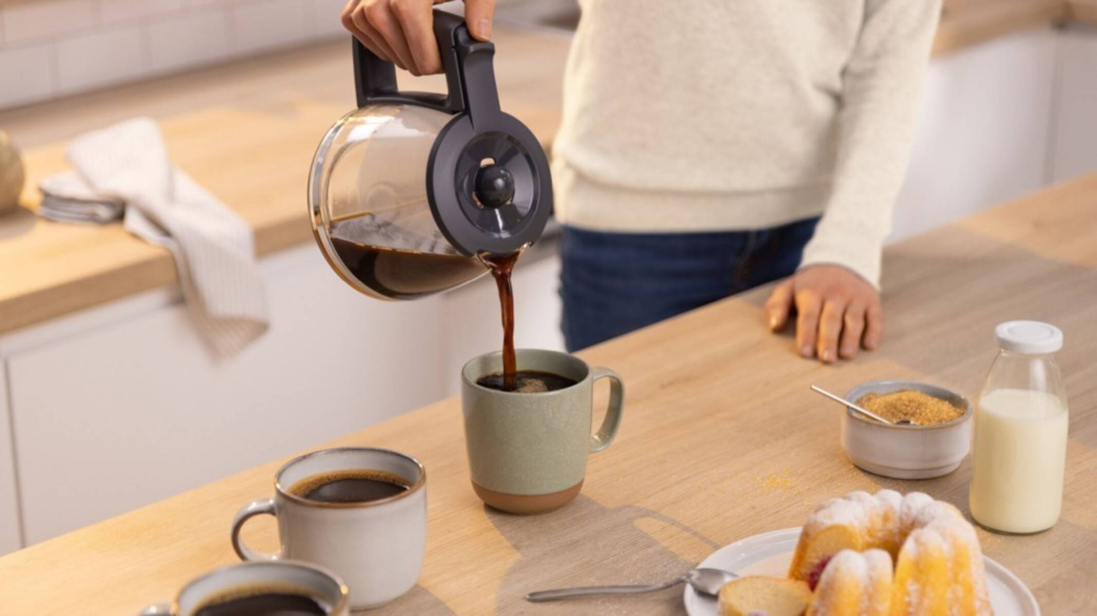 Eine Person gießt Kaffee aus einer Kanne in eine Tasse auf einer Küchenarbeitsplatte, wobei sich zwei weitere gefüllte Tassen in der Nähe befinden.