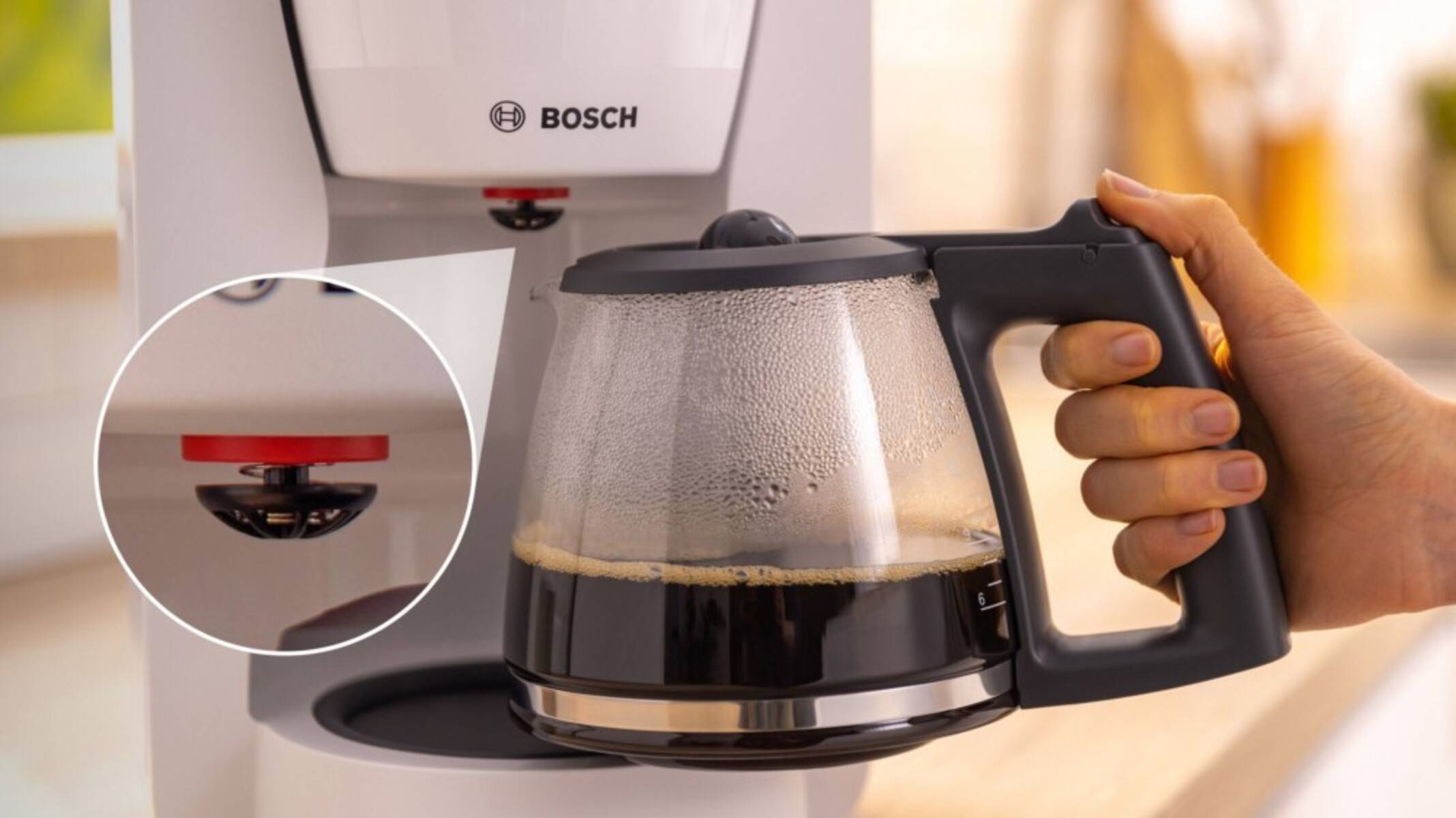 Eine Hand, die eine Glaskaffeekanne mit gebrühtem Kaffee hält, wobei der Bosch-Kaffeevollautomat von oben zu sehen ist. Ein Einsatz hebt einen roten Sicherheitsknopf hervor.