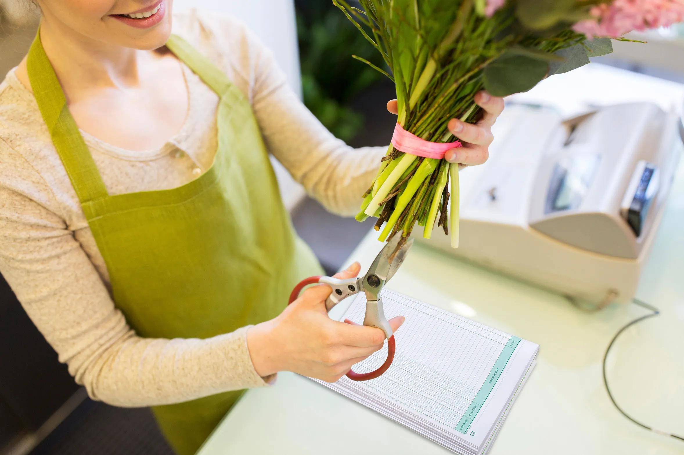 Ein Florist in einer grünen Schürze schneidet Blumenstiele mit einer Schere zurecht und lächelt dabei, während er an einem Tresen steht, neben dem ein offenes Auftragsbuch liegt.