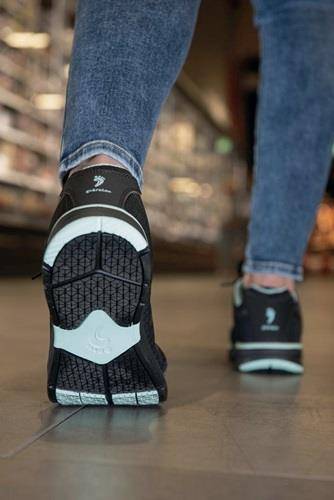 Eine Person in Jeans geht durch einen Ladengang und zeigt die Sohle eines Sneakers, der für Komfort konzipiert ist. Die Regale im Hintergrund sind unscharf.