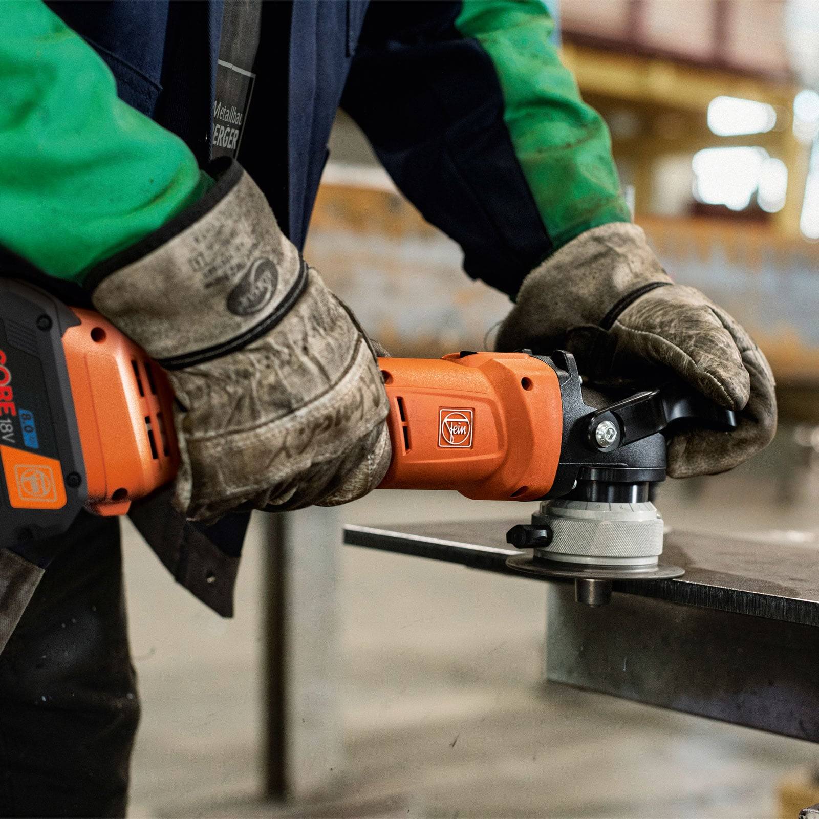 Eine Person mit Handschuhen benutzt eine Winkelschleifer auf einer Metalloberfläche in einer Werkstatt. Das Gerät ist orange und hat sichtbare Markierungen der Marke.