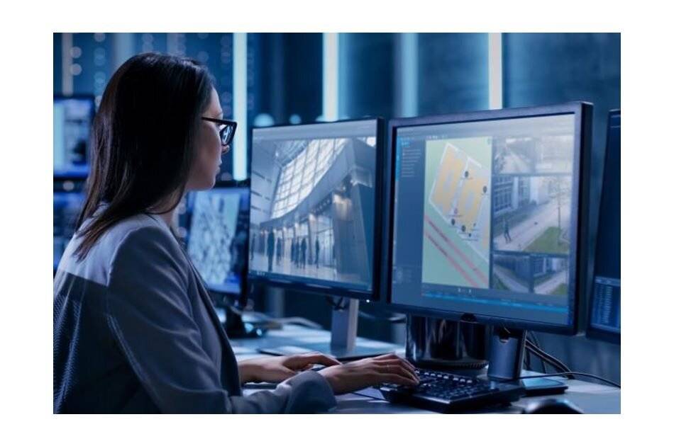 Eine Frau überwacht Sicherheitskameraaufnahmen und einen Gebäudeplan auf zwei Computerbildschirmen in einem schwach beleuchteten Kontrollraum.