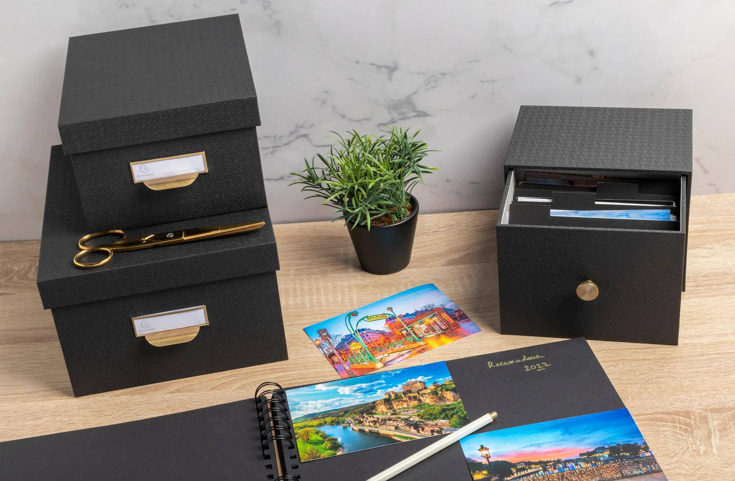 Ein aufgeräumter Schreibtisch mit schwarzen Aufbewahrungsboxen, einer kleinen Topfpflanze, einem leeren Notizblock und farbenfrohen Fotografien, was Ordnung und Kreativität andeutet.