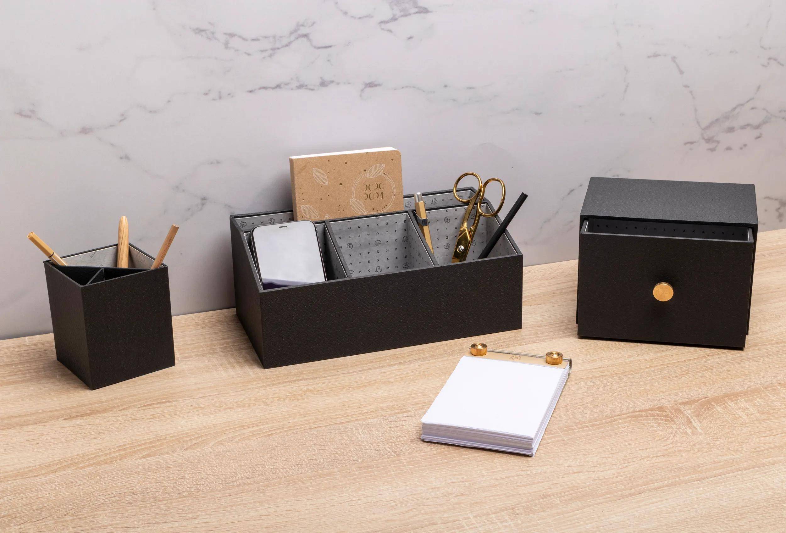 Schwarze Schreibtischorganizer auf einer Holzoberfläche enthalten Notizbücher, Stifte, Schere und ein Smartphone. Ein Stapel weißer Notizblöcke befindet sich in der Nähe.