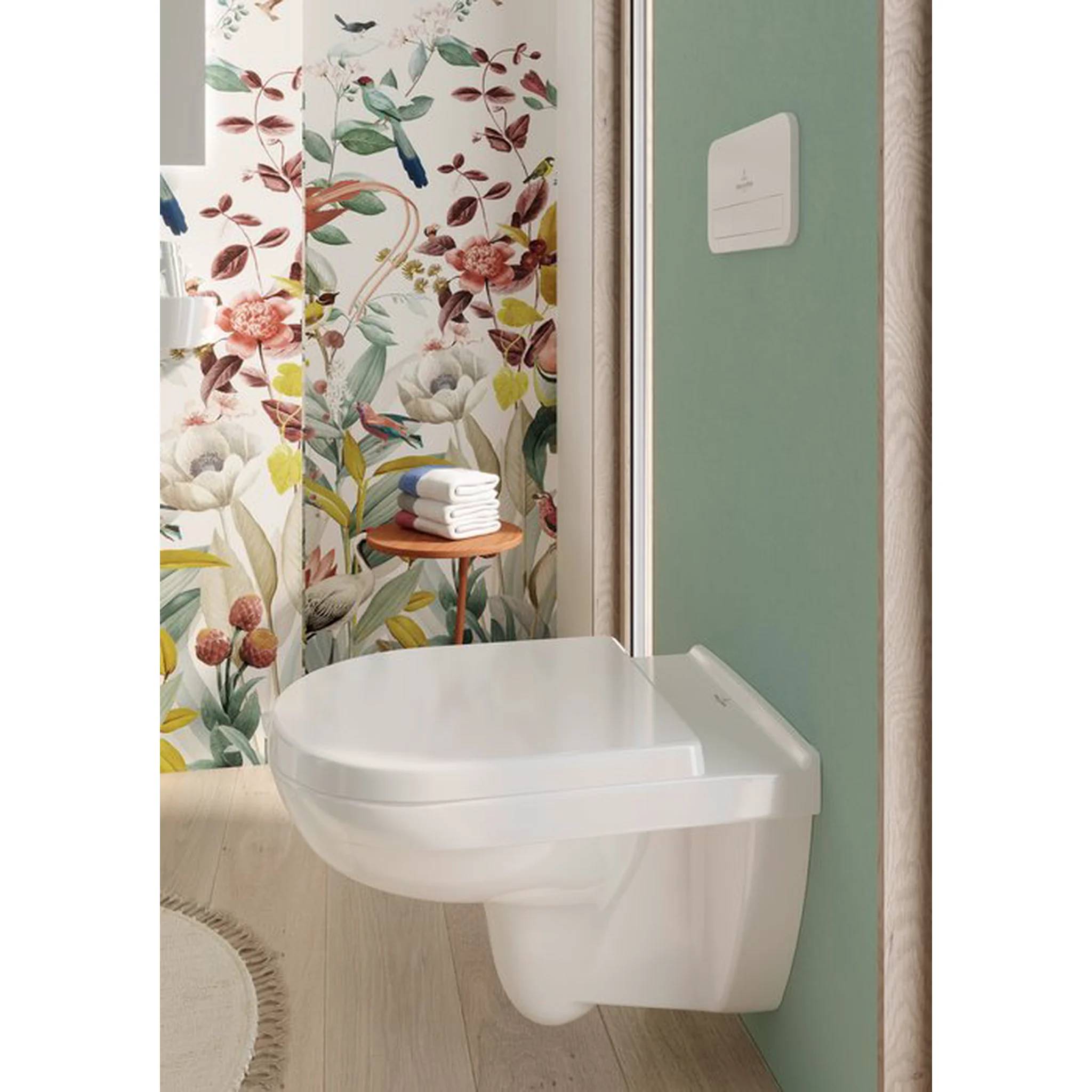 Weiße wandmontierte Toilette in einem Badezimmer mit mintgrünen Wänden und einem Duschvorhang mit Blumenmuster, mit gefalteten Handtüchern auf einem kleinen Beistelltisch.