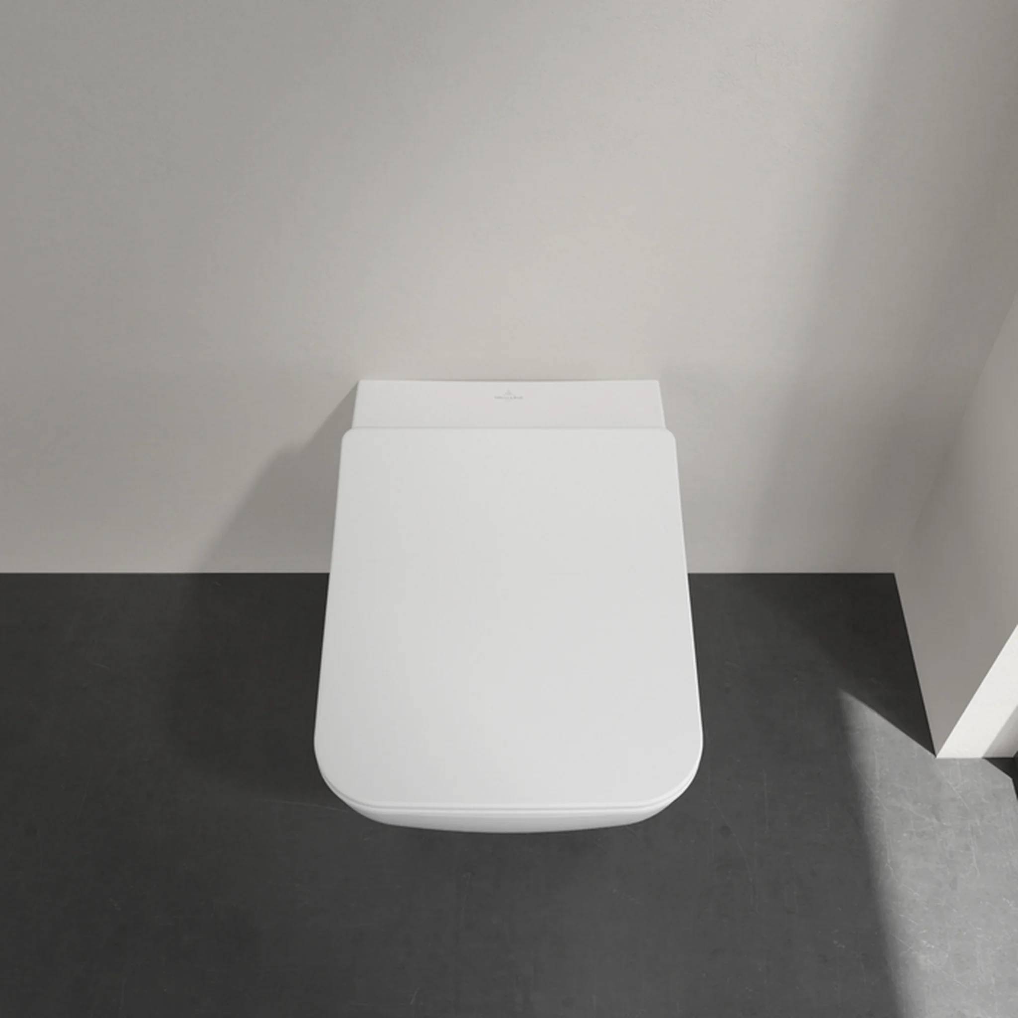 Weiße an der Wand montierte Toilette in einem minimalistischen Badezimmer, von oben betrachtet auf dunklen Bodenfliesen.