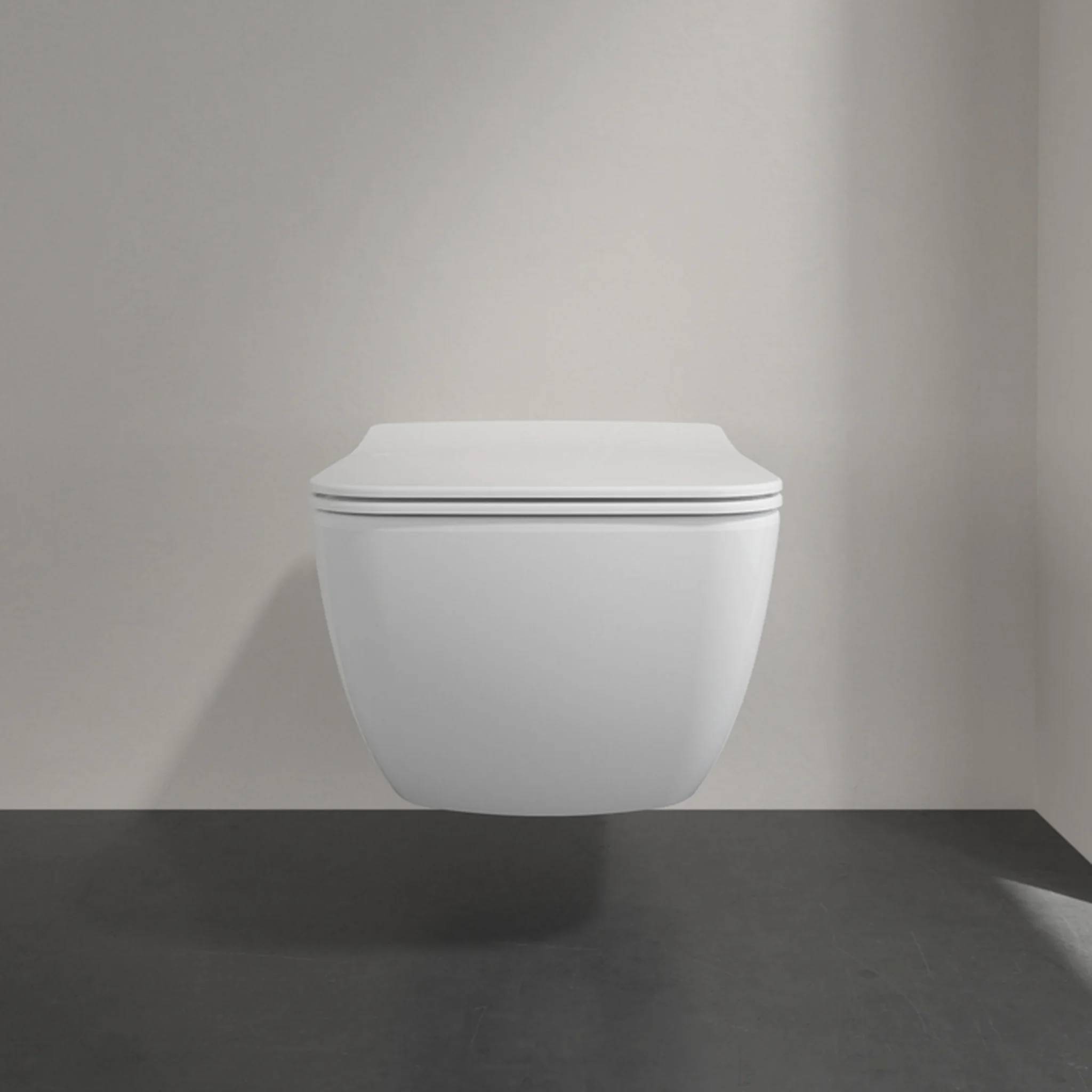 Modernes weiß montiertes Wandtoilette mit geschlossenem Deckel gegen eine schlichte graue Badezimmerwand und dunklem Boden.