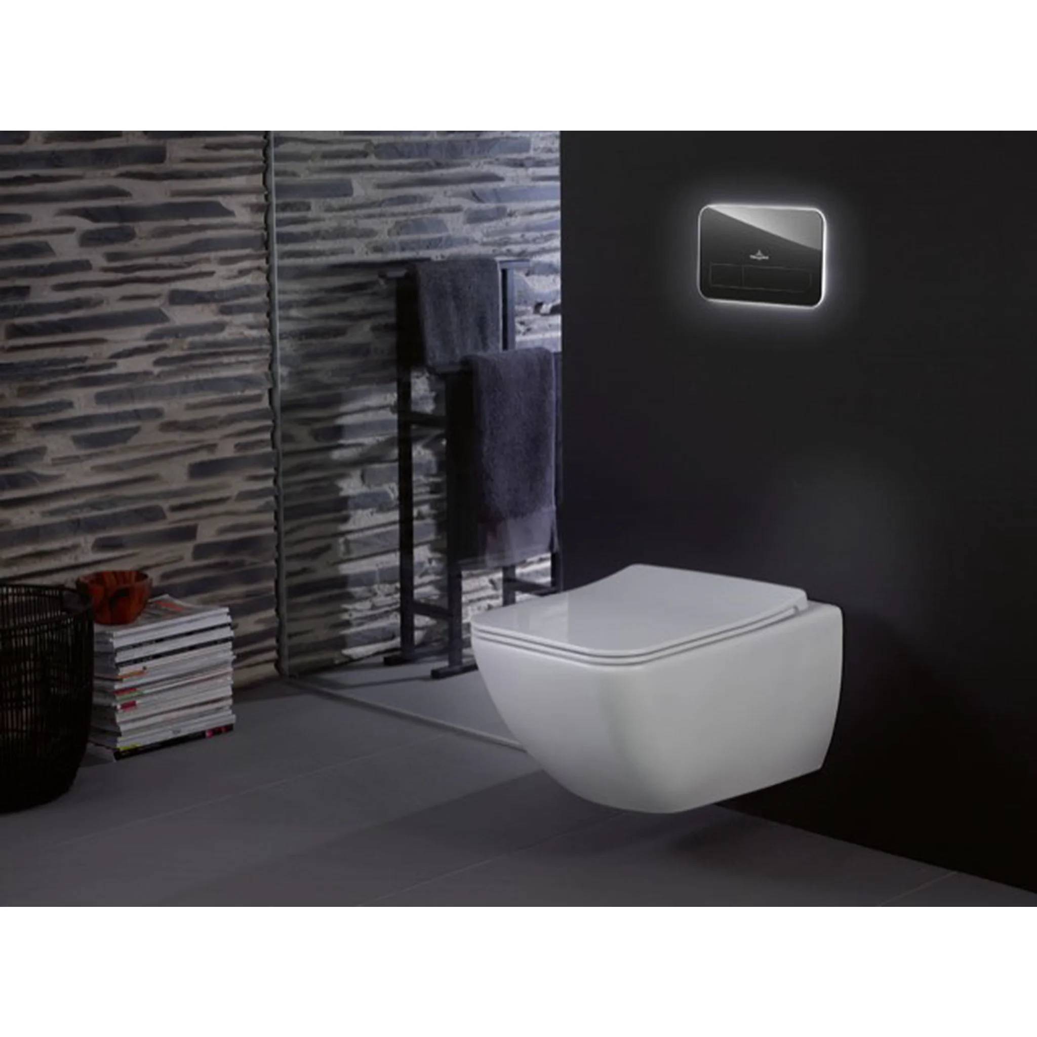 Modernes, an der Wand montiertes weißes WC in einem dunklen Badezimmer, mit einem leuchtenden Spülpaneel darüber und Handtüchern auf einem Handtuchständer in der Nähe.