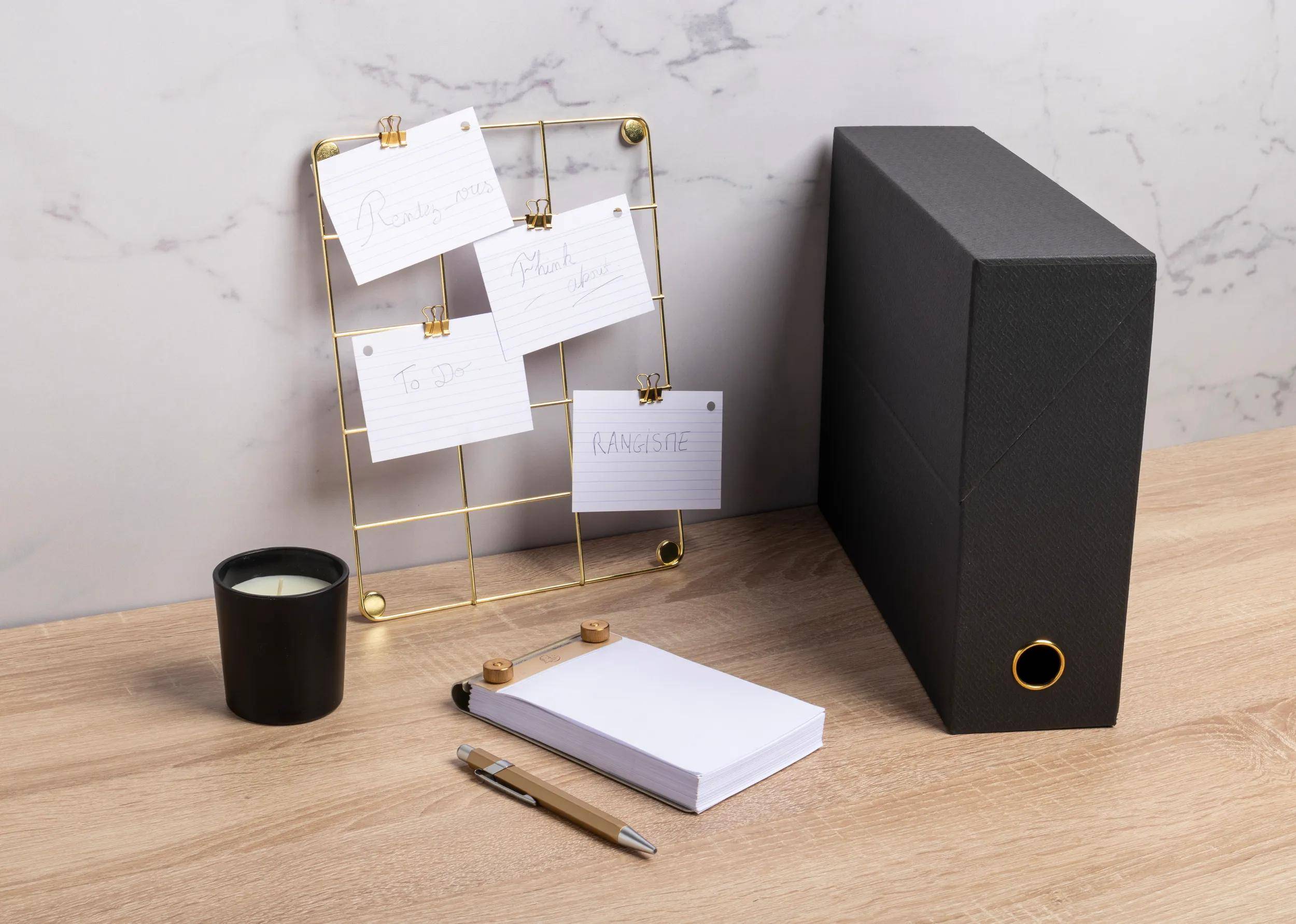 Eine Schreibtischeinrichtung mit einem schwarzen Dateiorganizer, einer Kerze, Stiften, einem Notizbuch und einem goldfarbenen Draht-Notizhalter, der kleine Karten auf einer Holzoberfläche anzeigt.