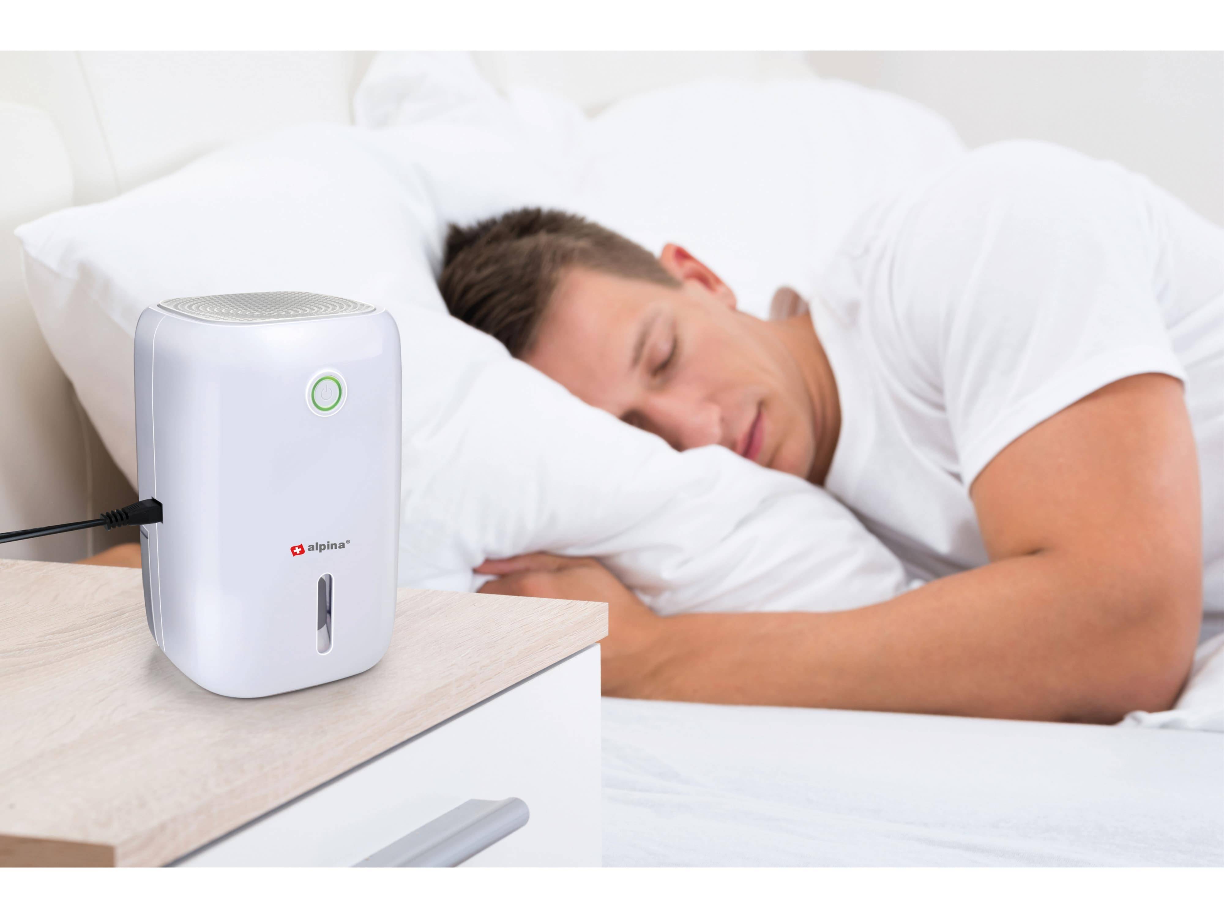 Eine Person schläft auf einem Bett mit einem kleinen weißen Luftreiniger auf einem Nachttisch in der Nähe, was auf einen Fokus auf saubere Luft während des Ausruhens hindeutet.