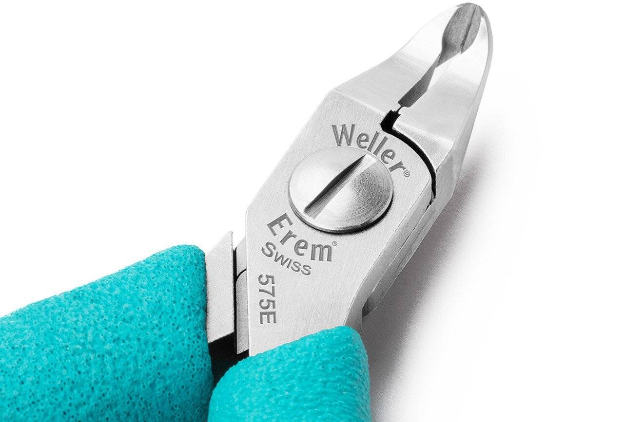 Nahaufnahme von türkisfarbenen behandschuhten Fingern, die eine silberne Weller Erem Swiss 575E Präzisionszange auf weißem Hintergrund halten.