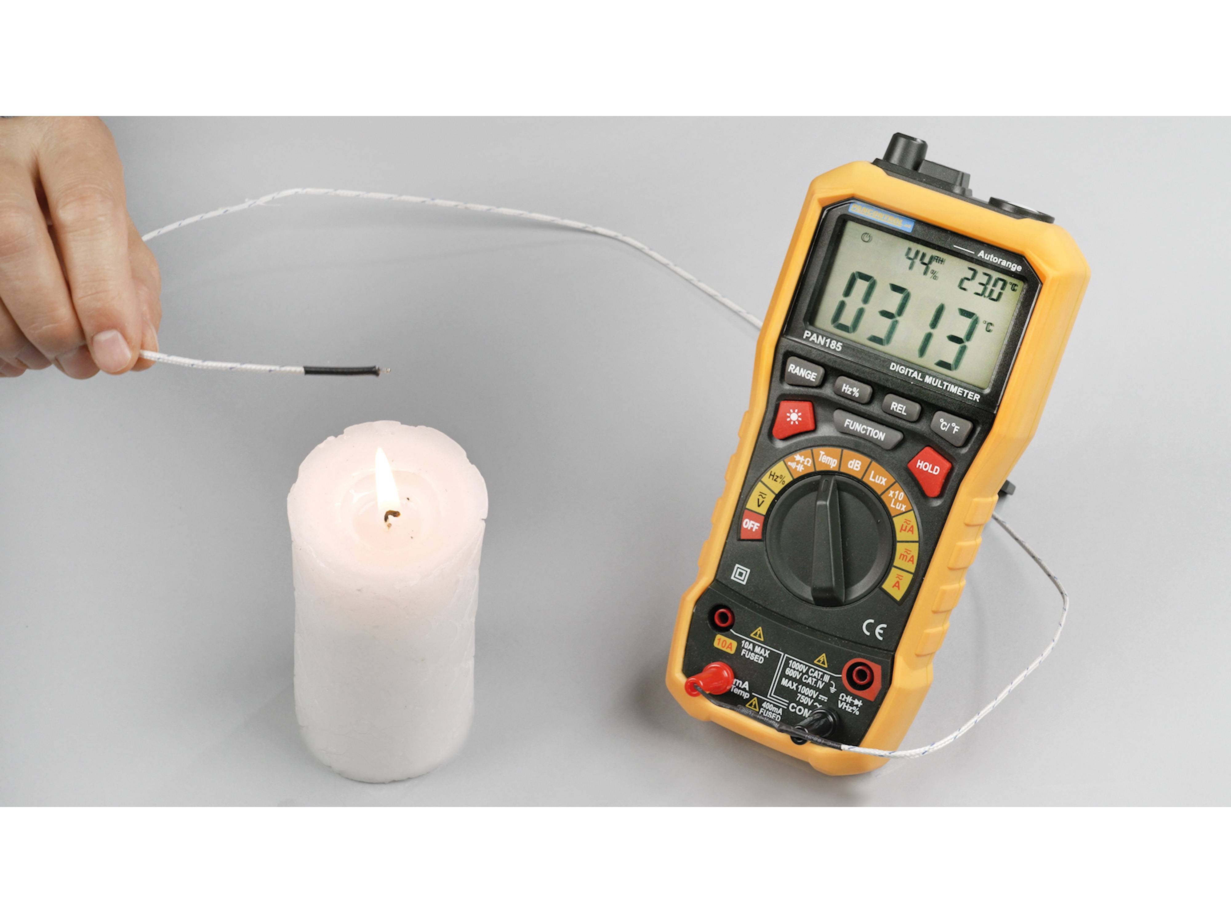 Ein digitales Multimeter mit gelbem Gehäuse misst die Spannung in der Nähe einer brennenden weißen Kerze und zeigt 
