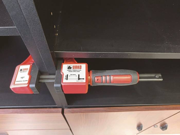 Eine Klemme mit der Aufschrift 'Urko Tools, S.A.' befestigt zwei dunkle Regale miteinander. Der Klemmgriff ist rot und schwarz und horizontal ausgerichtet.