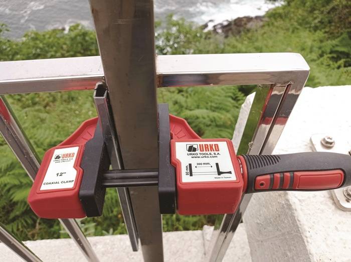 Ein Metallhandlauf, der mit einer roten Urko-Klemme installiert wird, mit Blick auf eine üppige grüne Landschaft und einem Gewässer im Hintergrund.