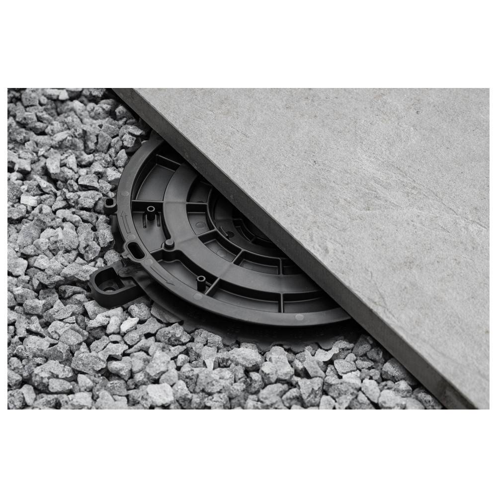 Ein schwarzes Deck-Stützelement, das teilweise von einer quadratischen grauen Fliese bedeckt ist, sitzt auf einem Bett aus grauem Kies und veranschaulicht eine Konstruktionsmethode.