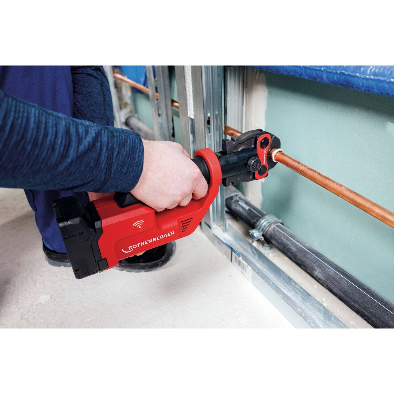 Eine Person verwendet ein rotes Rohrabschneider-Werkzeug, um ein Kupferrohr in einem Baustellenumfeld zu schneiden. Das Werkzeug wird sicher gehalten, und das Rohr ist im Schneider positioniert.