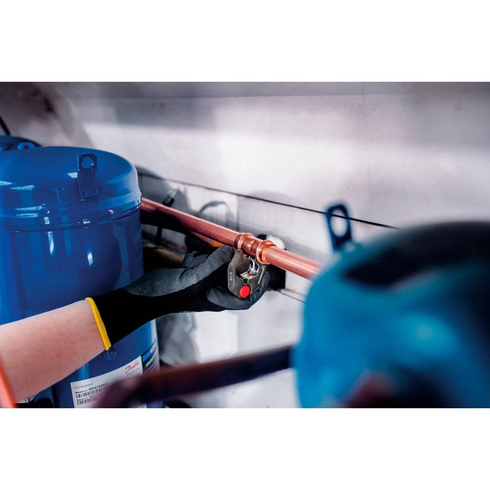 Eine Person mit Handschuhen verwendet einen Schraubenschlüssel, um ein Ventil an einem Kupferrohr neben einem blauen Tank zu festigen, was Wartungsarbeiten andeutet.