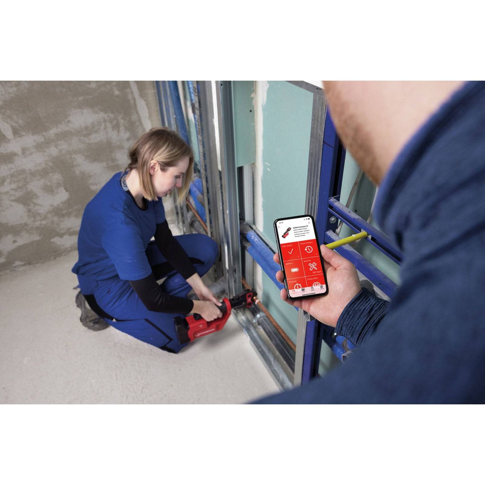 Eine Person in blauer Arbeitskleidung benutzt ein Werkzeug an einem Metallrahmen. Eine andere Person hält ein Smartphone, das eine App mit einer roten Benutzeroberfläche zeigt.