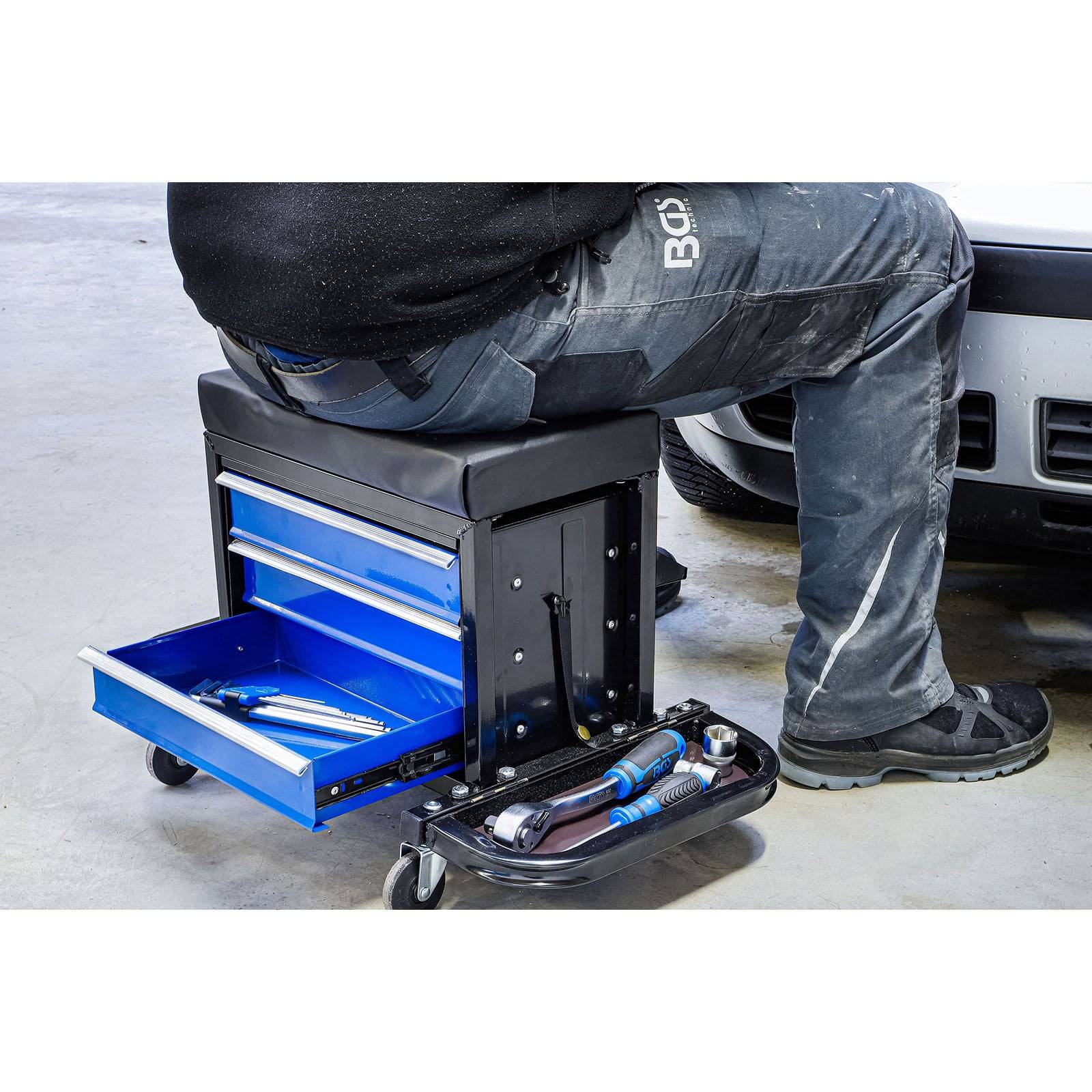 Eine Person sitzt auf einem fahrbaren Werkzeugschrank neben einem Auto. Der Schrank hat offene Schubladen mit Werkzeugen, was auf eine Reparatur- oder Wartungsaufgabe hindeutet.