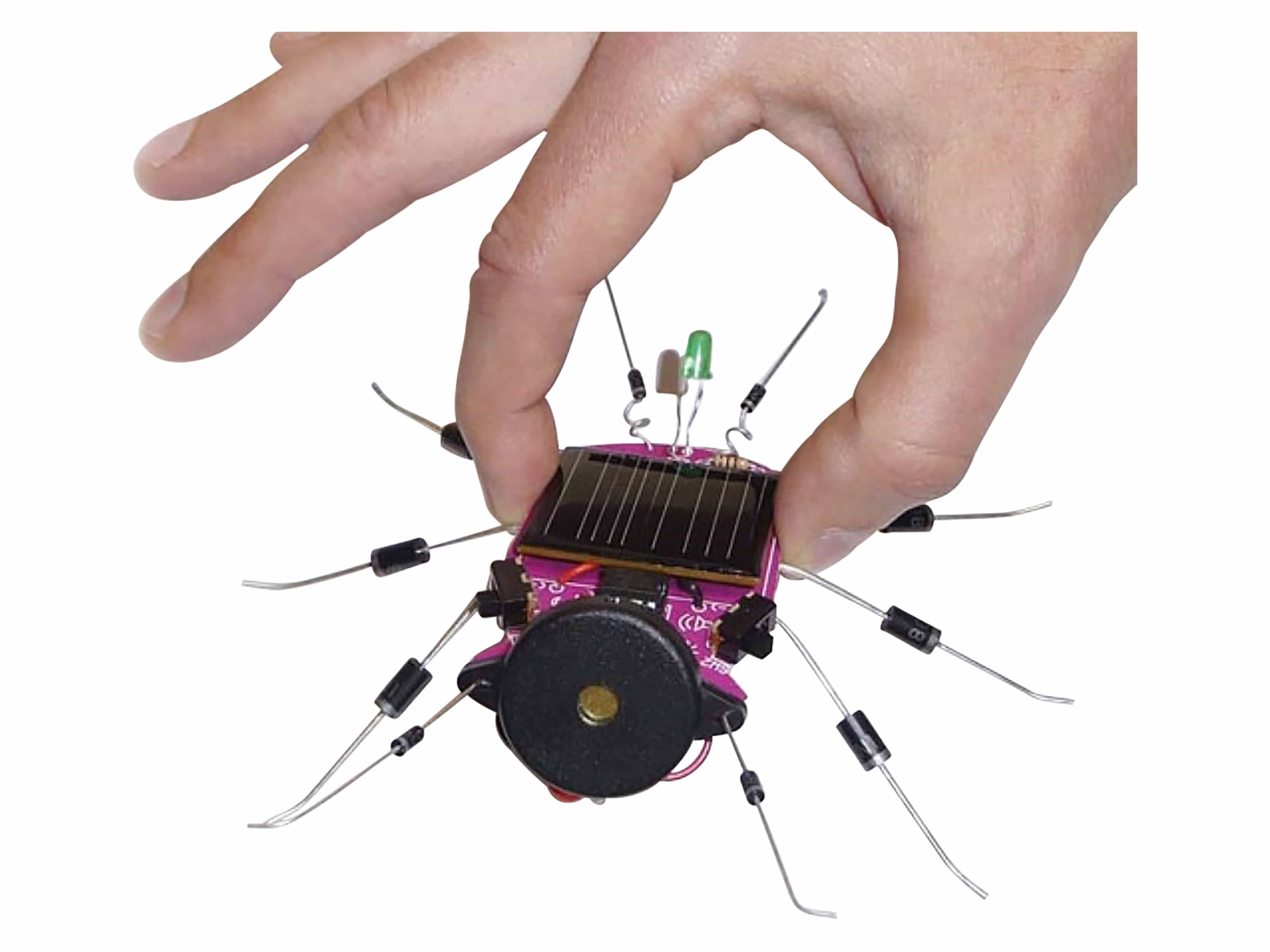 Eine Hand greift nach einem kleinen, solarbetriebenen, insektenähnlichen Roboter mit Drahtbeinen und rundem Körper, was eine spielerische Interaktion mit Technologie veranschaulicht.