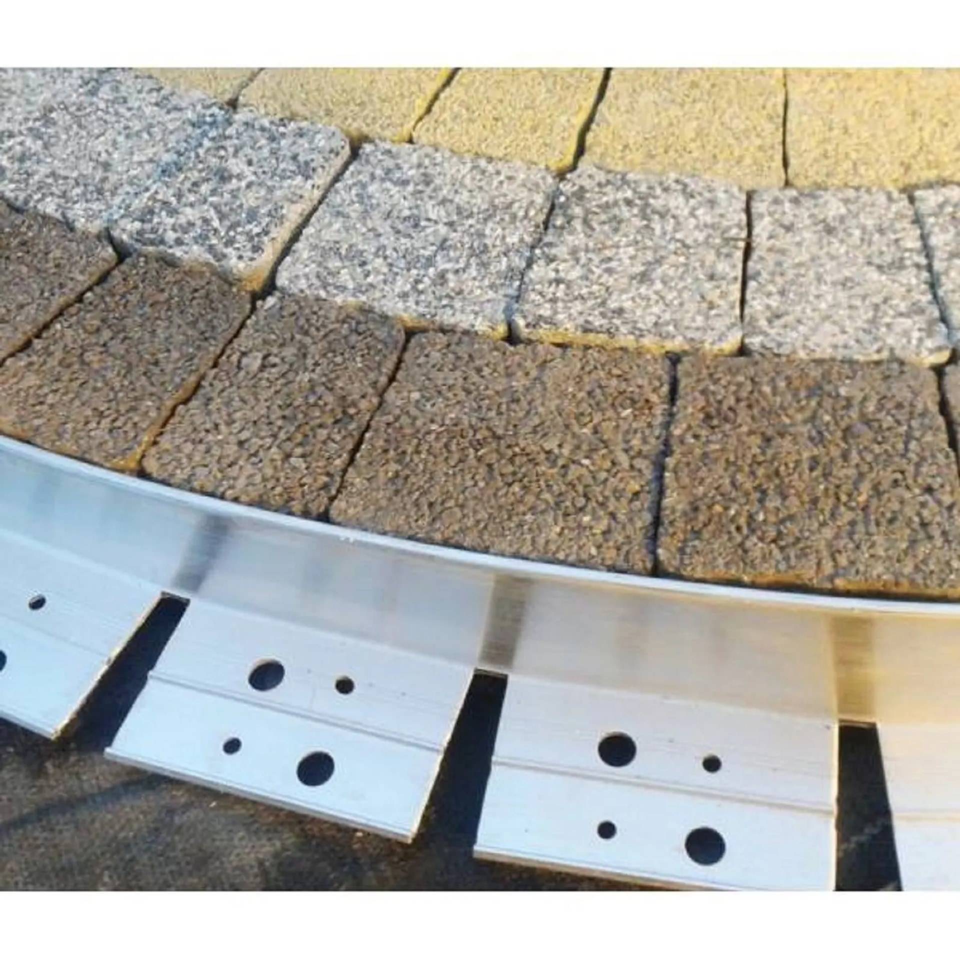 Nahaufnahme von geschichteten Pflastermaterialien und Einfassungen: gebogene Reihen von gelben, grauen und braunen Zuschlagsteinen über einem silbernen Metallhaltestreifen mit Schraubenlöchern.